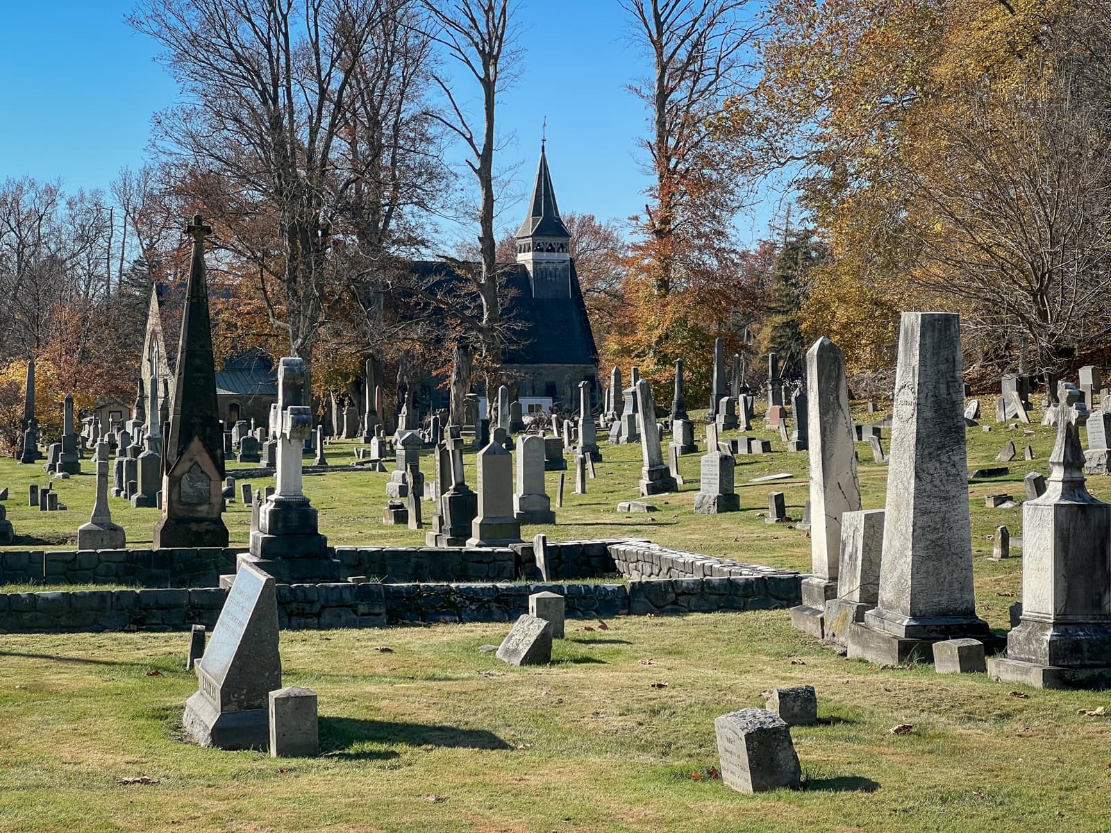 This image depicts a peaceful cemetery scene, with a variety of gravestones and monuments set against a backdrop of tall trees and a quaint chapel or church. The stones range in size and shape, from simple markers to tall obelisks, creating a sense of history and reverence. The bright blue sky and autumnal trees in the background, with their golden and orange leaves, add warmth to the otherwise solemn setting. The overall mood is serene, evoking reflection and calmness, as the landscape balances the natural beauty of the surroundings with the stillness of the graveyard.