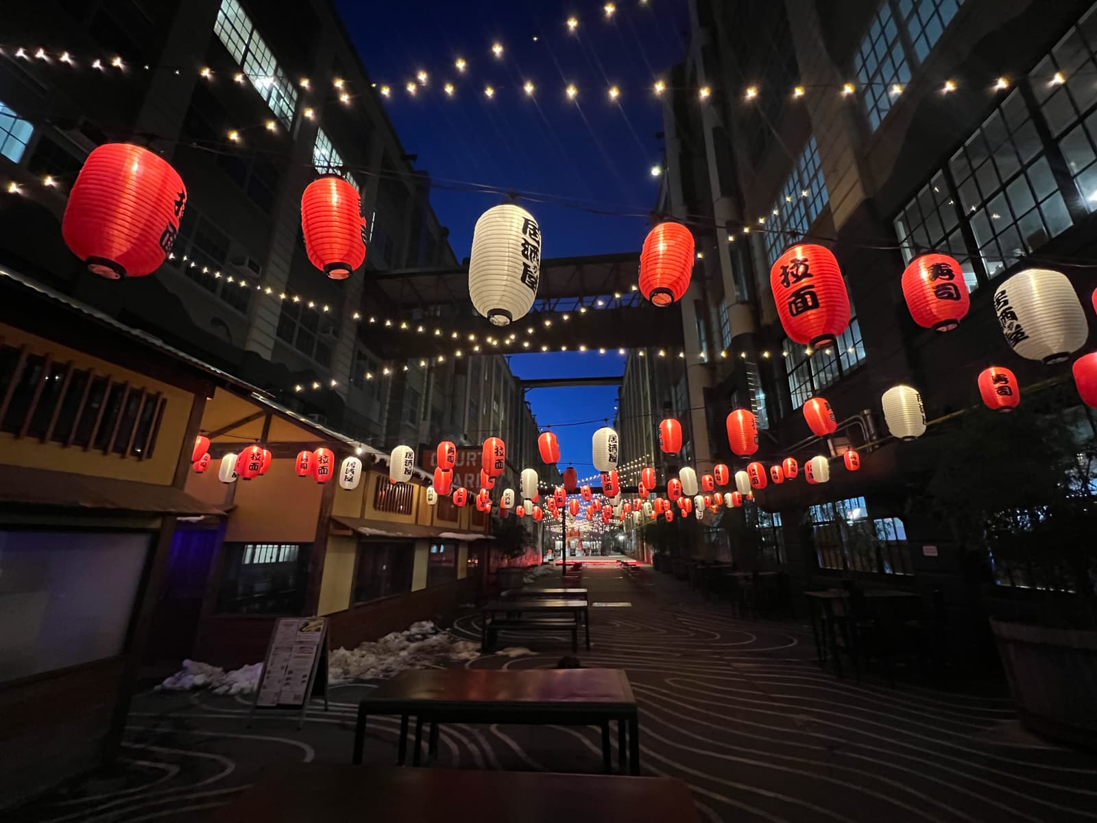 A narrow urban alleyway adorned with strings of red and white lanterns glowing softly in the evening. The lanterns, hanging in rows across the street, create a festive and lively atmosphere as the lights twinkle against the backdrop of tall buildings on either side.