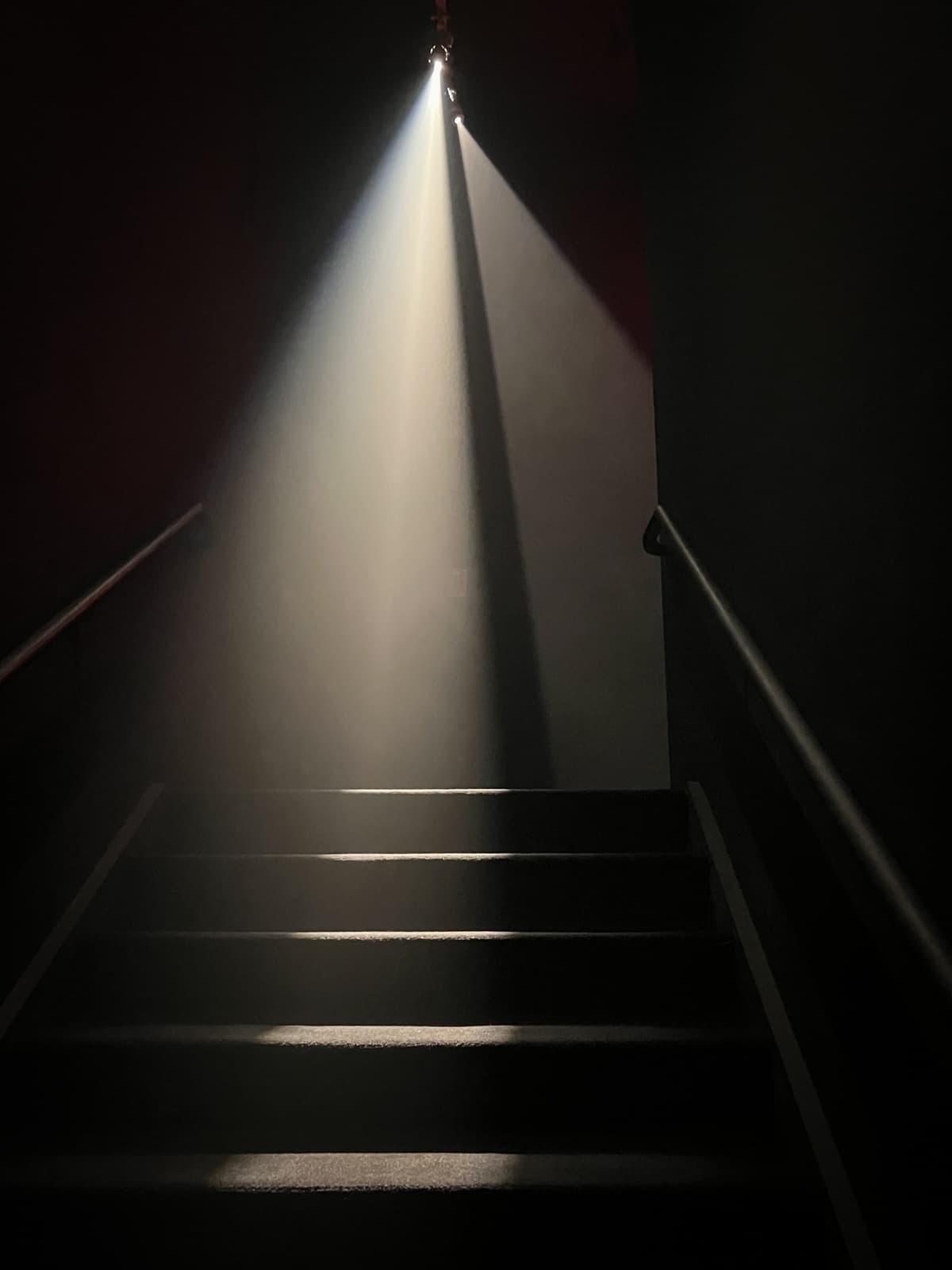 A dark staircase illuminated by a single bright beam of light coming from above, casting sharp shadows and creating an ethereal, dramatic effect. The focus is on the contrasting light and shadow, emphasizing the texture and depth of the scene.