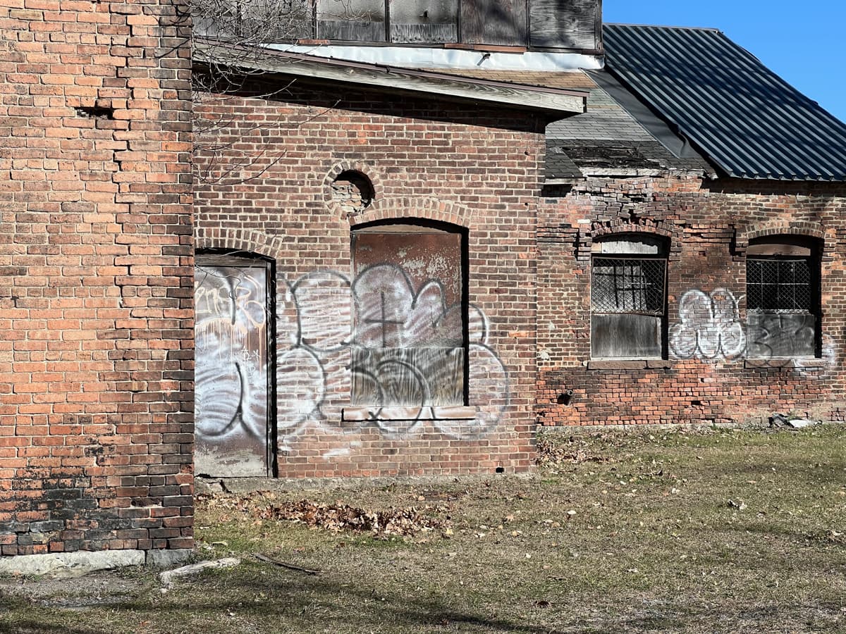 An old brick building with boarded-up and broken windows, covered in graffiti. The structure shows signs of decay, with crumbling bricks and weathered surfaces. The ground is bare with patches of grass and fallen leaves, suggesting it is in an abandoned or neglected state.