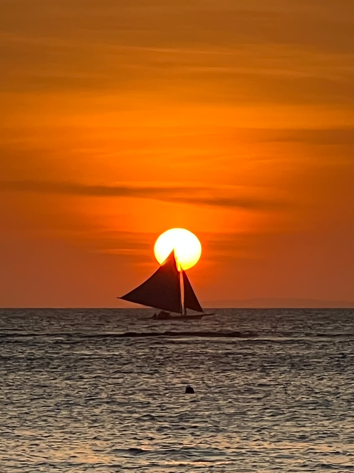 A sailboat silhouetted against a vibrant orange sunset, with the sun perfectly positioned behind its sail as it glides across calm ocean waters.