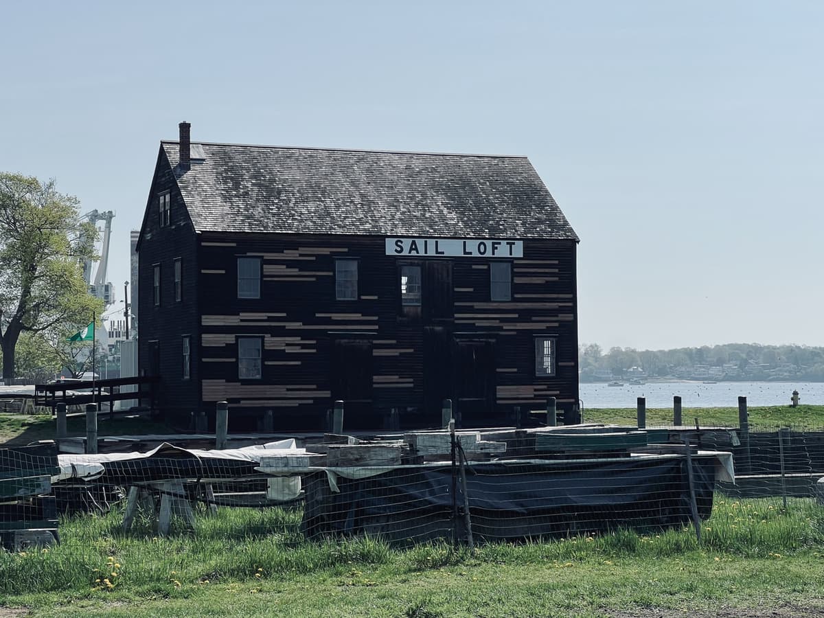Historic wooden building labeled ‘Sail Loft’ near a waterfront, with scattered planks and materials in the foreground and a calm body of water behind it.