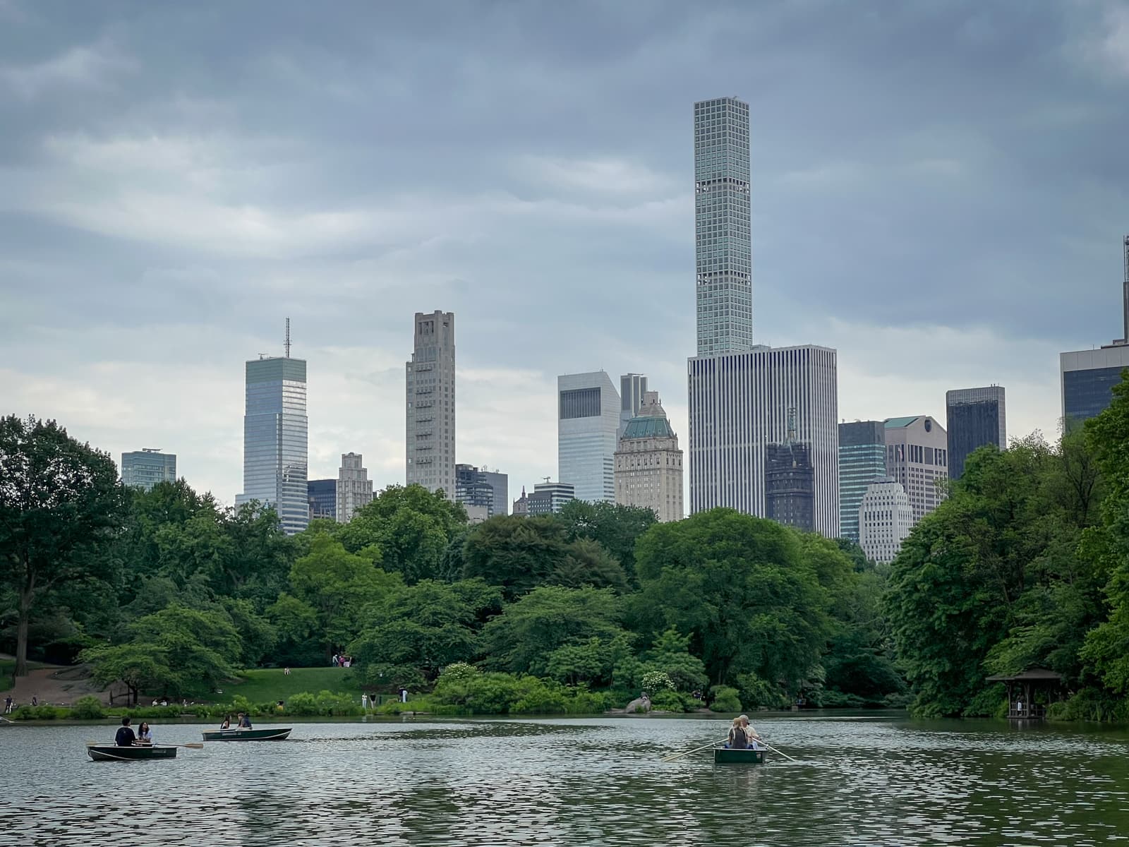 View of Central Park Lake with people rowing boats in the foreground, and the Manhattan skyline rising in the background under an overcast sky.