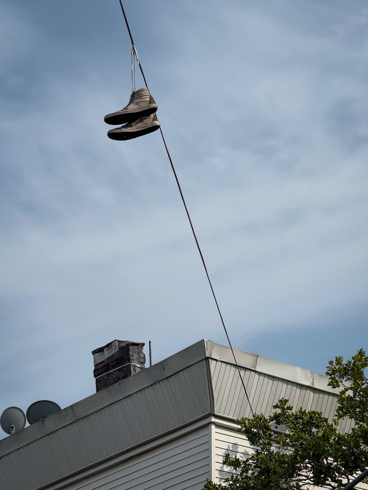 A pair of sneakers hanging from a wire against a cloudy sky, above a building with white siding, a chimney, satellite dishes, and part of a tree visible.