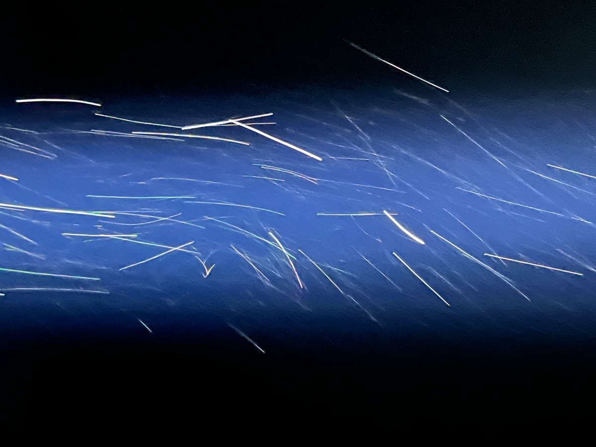 A photograph capturing streaks of light against a dark blue background, with thin, bright lines cutting across the frame diagonally. The image evokes a sense of motion, resembling sparks or light trails moving swiftly through space, creating a feeling of energy and speed.