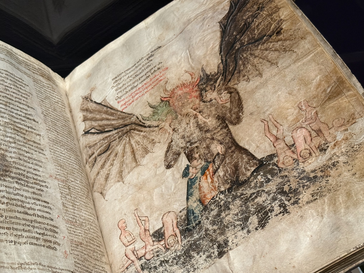 An open medieval manuscript displaying a full-page illuminated illustration depicting Satan in Hell, consistent with imagery from Dante's Inferno. The central figure is a enormous winged demon with multiple heads and bat-like wings, surrounded by small naked human figures representing the damned. The illustration is rendered in muted earth tones — browns, pinks, and greens — with visible age, foxing, and wear across the vellum. Red and black handwritten text in Italian appears above the illustration and in columns on the facing left page.