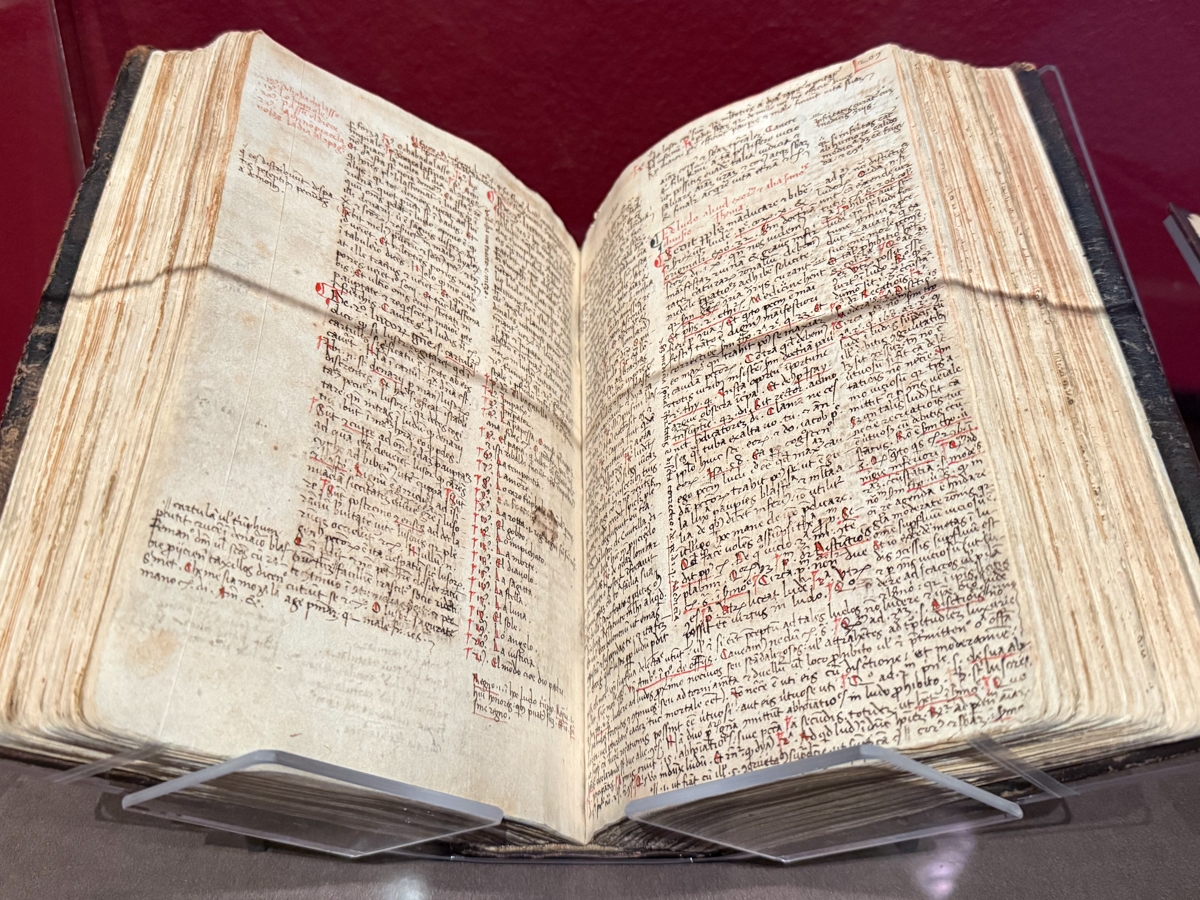 An open medieval manuscript displayed on a clear acrylic book stand against a deep red background. The thick, aged volume lies open to a two-page spread densely covered in small handwritten Latin script in brown ink, with red ink used for headings, annotations, and marginal notes throughout. The pages are yellowed and worn with age, and the dark leather binding is visibly weathered.