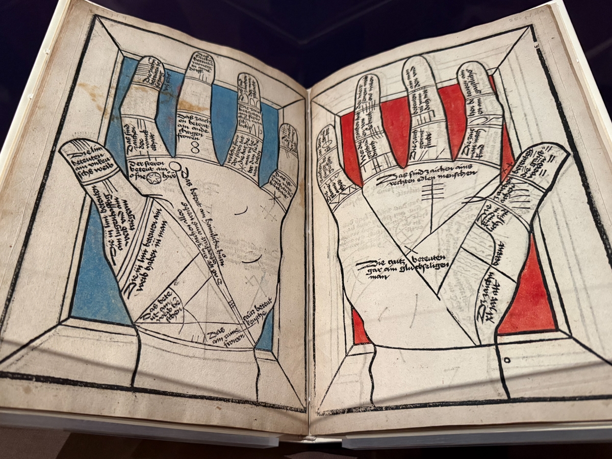An open early printed book displayed under dark lighting, showing a two-page spread with woodcut diagrams of two outstretched hands viewed palm-up. The left hand is colored with blue-highlighted fingers and the right with red, each divided into labeled zones with German text identifying lines and regions used in palmistry. The illustrations are precise and diagrammatic, resembling a chiromancy or fortune-telling manual from the 15th or 16th century.