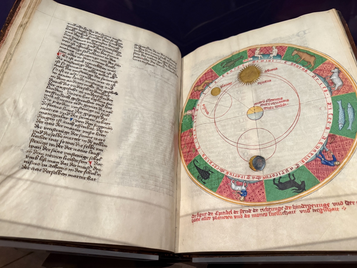 An open medieval manuscript showing a dense column of German Gothic script on the left page and a richly illuminated astronomical diagram on the right. The diagram depicts a geocentric model of the cosmos as a circular chart, with concentric rings labeled with planet names and zodiac signs. The outer border is decorated with alternating red and green hatched segments, each containing a painted zodiac animal. A gold sun and gray moon are positioned within the inner rings. A red caption in German runs below the illustration.