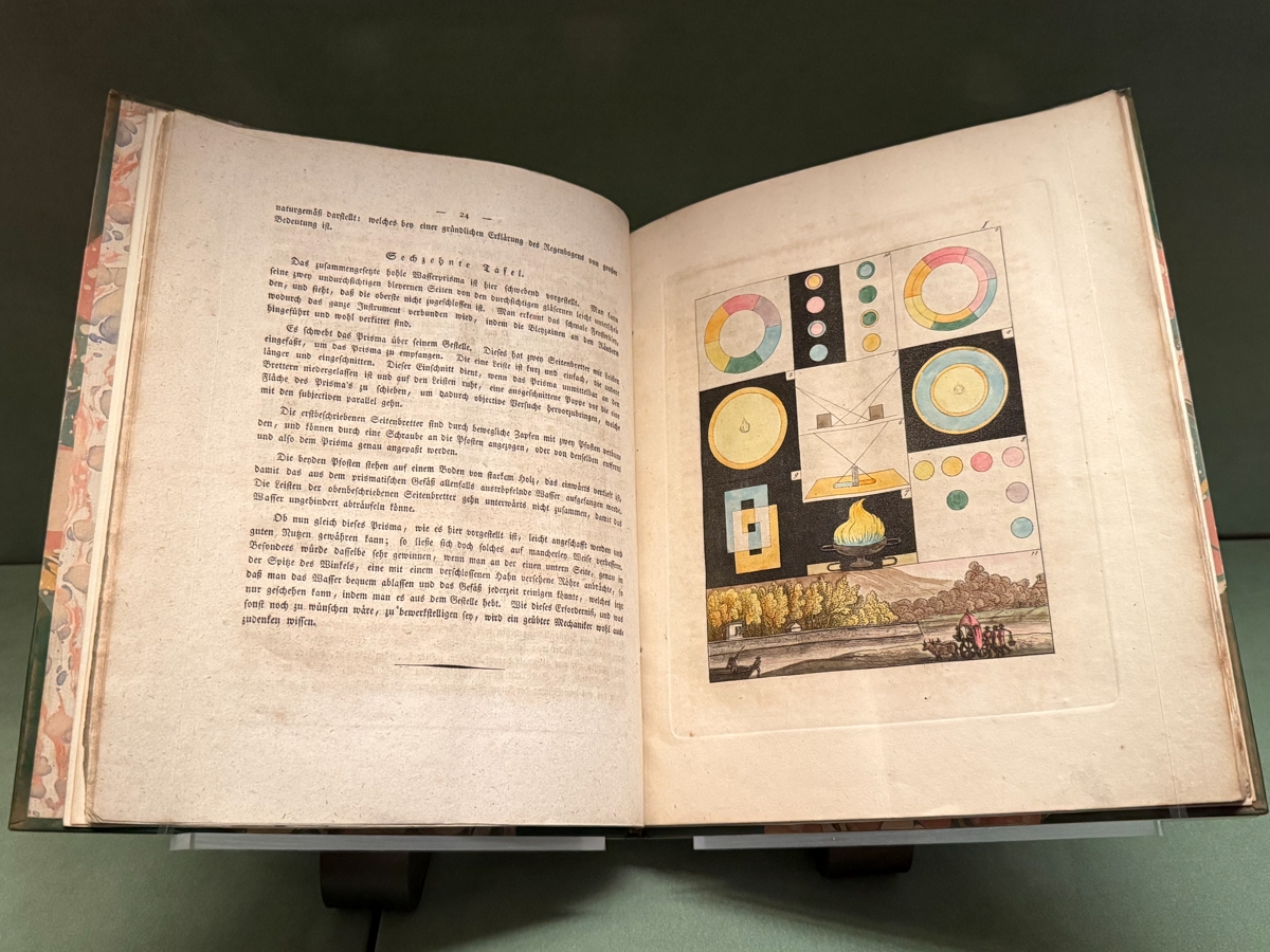 An open 18th or early 19th century German scientific book on a display stand against a gray-green background. The right page features a hand-colored engraved plate labeled Sechzehnte Tafel (Sixteenth Plate), showing optical experiments related to color and light — including color wheels, geometric diagrams, colored circles on black backgrounds, a prism refracting light, a flame, and a landscape scene. The left page contains typeset German text with a centered heading. The book has a decorative marbled cover.