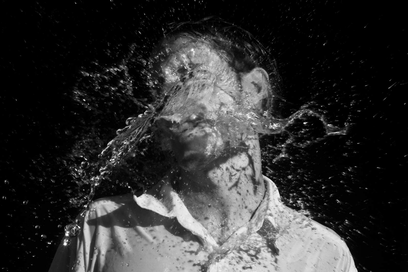 This black-and-white photograph captures a moment of impact as water splashes across a man's face, frozen in time. The water forms intricate patterns of droplets and waves, partially obscuring his face while creating a dynamic sense of movement. The man's expression is not fully visible due to the splash, but the action suggests a sudden, forceful dousing. The dark background contrasts sharply with the bright highlights of the water, emphasizing the texture and fluidity of the liquid. The image conveys a sense of shock or surprise, with the water suspended in mid-air adding a dramatic, almost surreal quality.