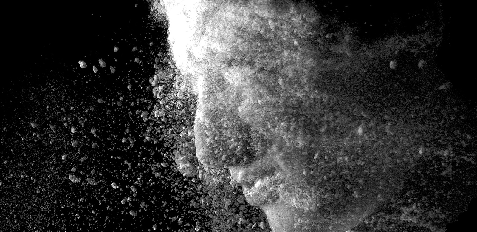 This black-and-white photograph captures a close-up side profile of a person, with their face partially obscured by what appears to be particles of dust or powder, as if they are dissolving or emerging from it. The fine particles scatter in different directions, giving the image a sense of motion and texture. The lighting highlights the contours of the face while emphasizing the granular quality of the dust. The dark background contrasts sharply with the illuminated face and particles, creating a dramatic and surreal effect, evoking themes of transformation, fragmentation, or disintegration.