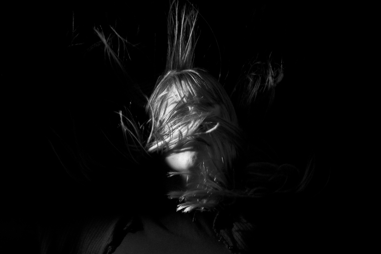 This black-and-white photograph shows a person with their face obscured by strands of hair, which are illuminated in sharp contrast against a dark background. The hair appears to be floating or blown by wind, adding a sense of motion and mystery to the image. Only part of the face is visible, emphasizing the eyes and nose, while the rest is hidden beneath the tangled hair. The play of light and shadow creates a dramatic, almost haunting atmosphere, as the figure seems to emerge from or be consumed by the darkness. The image evokes feelings of vulnerability, disorientation, or isolation.