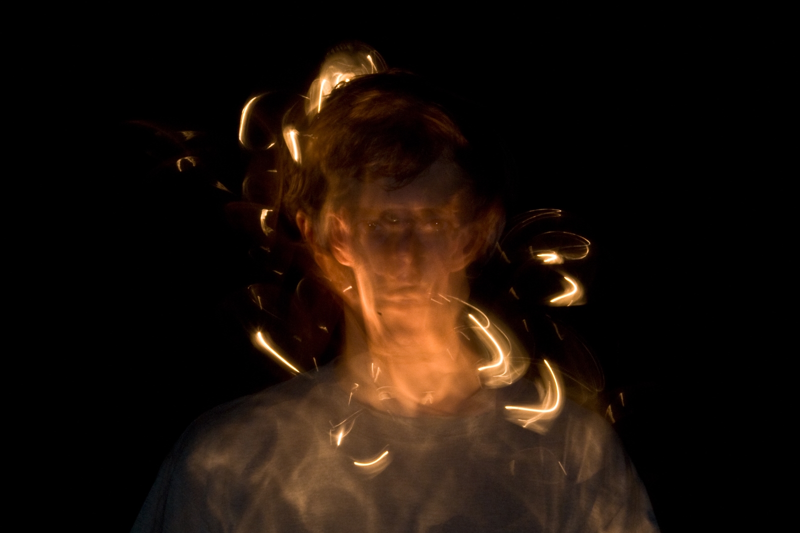This photograph features a long-exposure image of a person, creating a blurred and distorted effect that gives the figure a ghostly appearance. The person's face and body are partially transparent, with swirling streaks of warm light wrapping around them. The light trails, likely from moving sources, create a dynamic, abstract composition against the dark background. The combination of the subject's stillness and the fluid movement of light evokes a sense of surrealism and otherworldliness, as if capturing the essence of motion and time in a single moment. The image has an ethereal, dreamlike quality.
