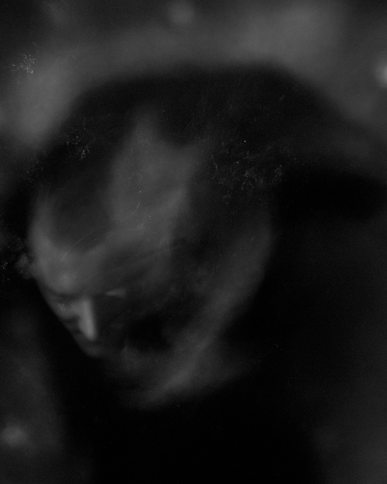 This black-and-white photograph features a blurred, ghostly image of a person’s face, partially obscured by swirling shadows and mist-like effects. The face appears faint and ethereal, blending into the dark background, which adds to the mysterious, dreamlike quality of the image. The soft focus and abstract nature create a sense of movement and introspection, as if the figure is emerging from or dissolving into the darkness. The overall atmosphere is haunting and otherworldly, evoking feelings of solitude and reflection.