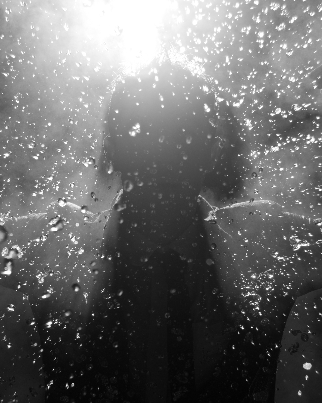 This black-and-white photograph captures a silhouette of a person behind a surface covered in water droplets. The light source, positioned directly above the figure, creates a dramatic backlit effect, causing the droplets to glisten and the person's form to appear shadowed and indistinct. The water droplets scatter the light, adding a sense of texture and depth to the image, while the overall mood feels mysterious and ethereal. The person’s arms are outstretched, creating a subtle, ghostly outline through the misty and watery atmosphere.
