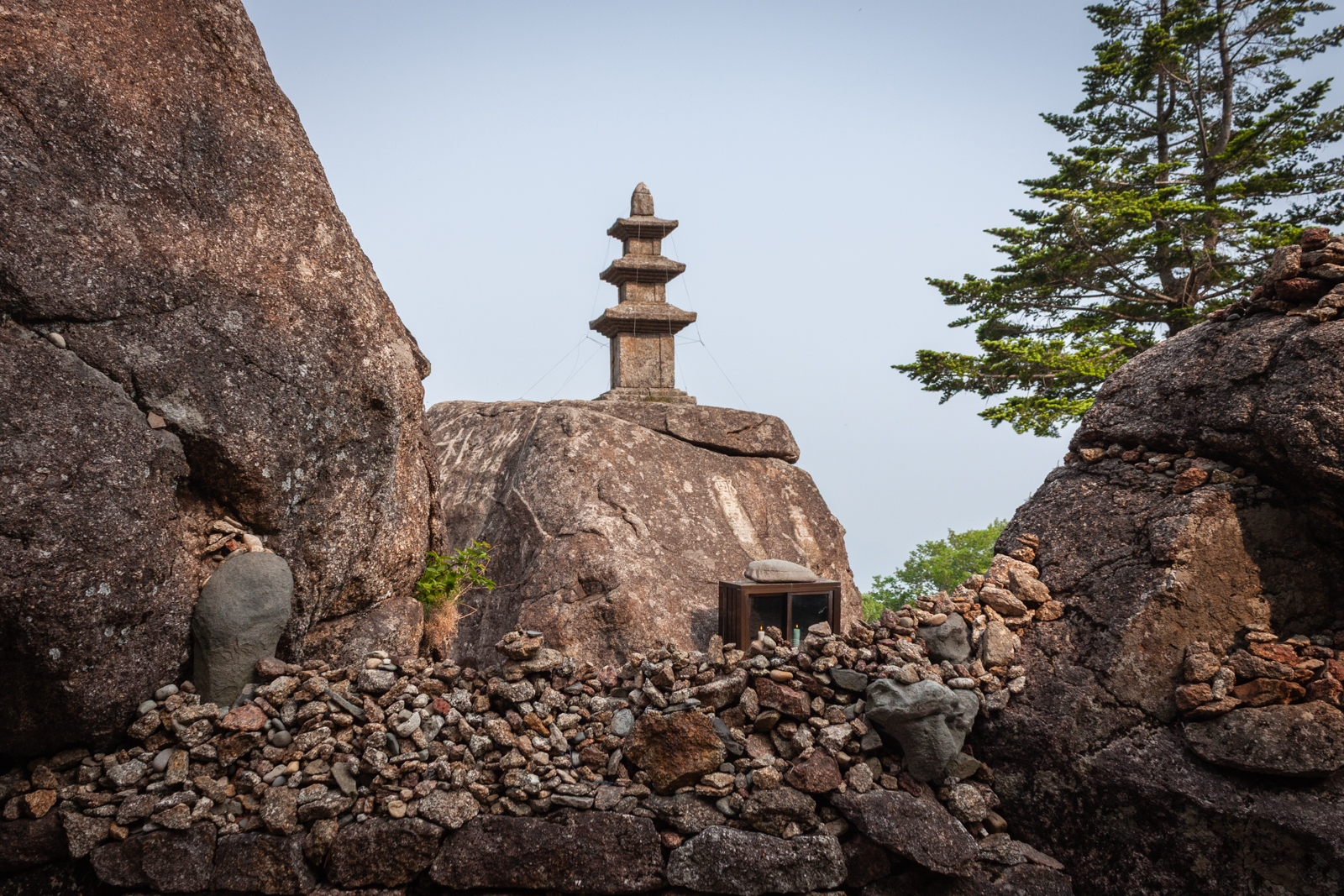 A stone pagoda perched on a large boulder surrounded by rocks and small stacked cairns, with a glimpse of greenery in the background.