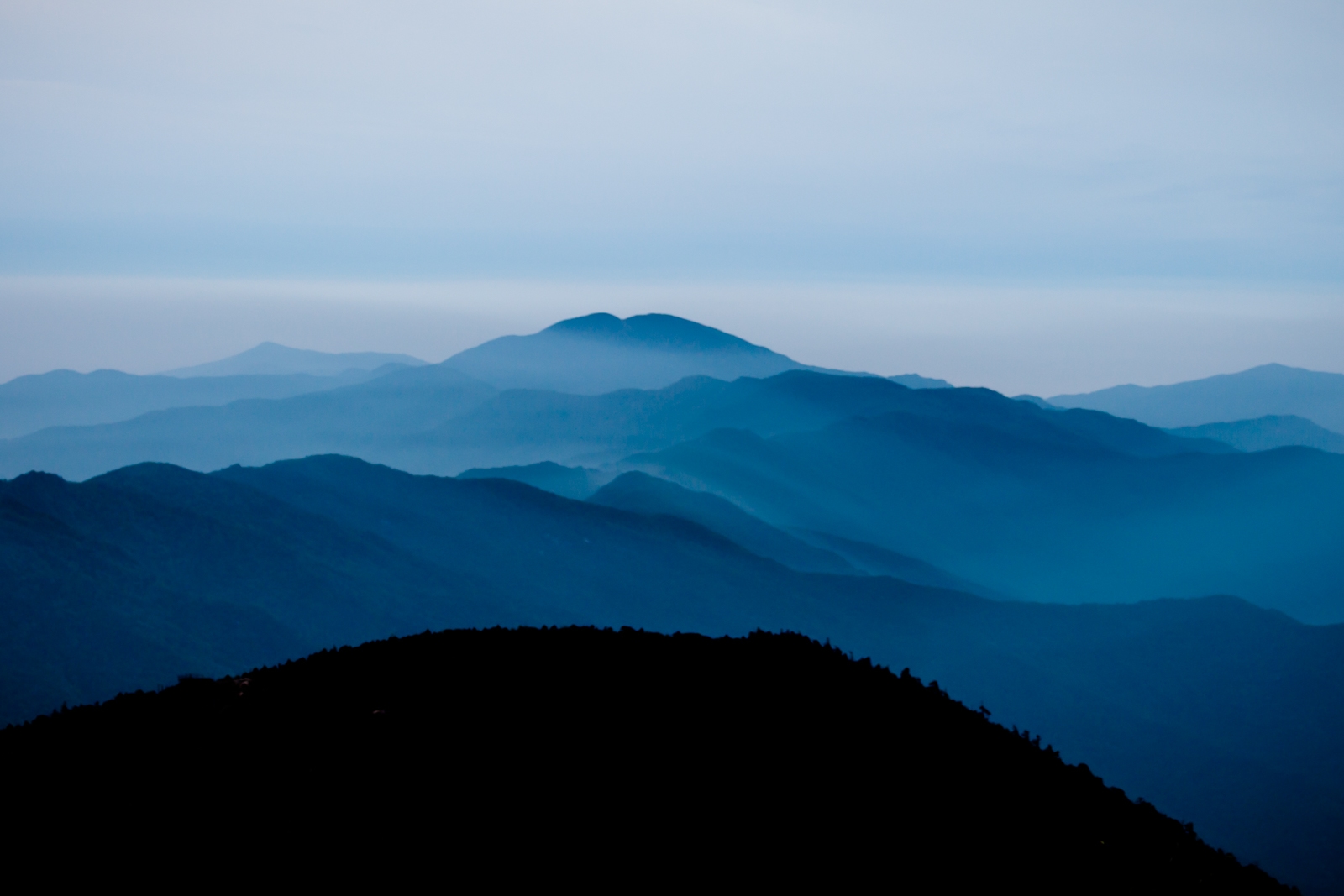 A serene view of blue-tinged mountain ranges fading into the misty distance, with gentle layers of hills and valleys stretching toward the horizon.
