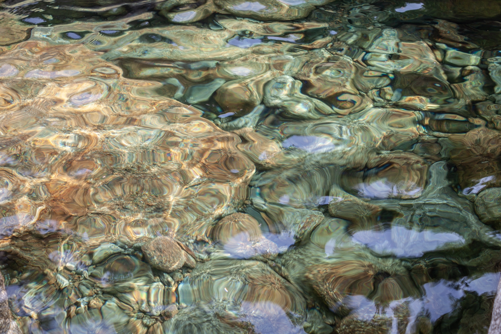 A close-up view of rippling water over colorful rocks, with the water surface distorting the shapes and colors below.