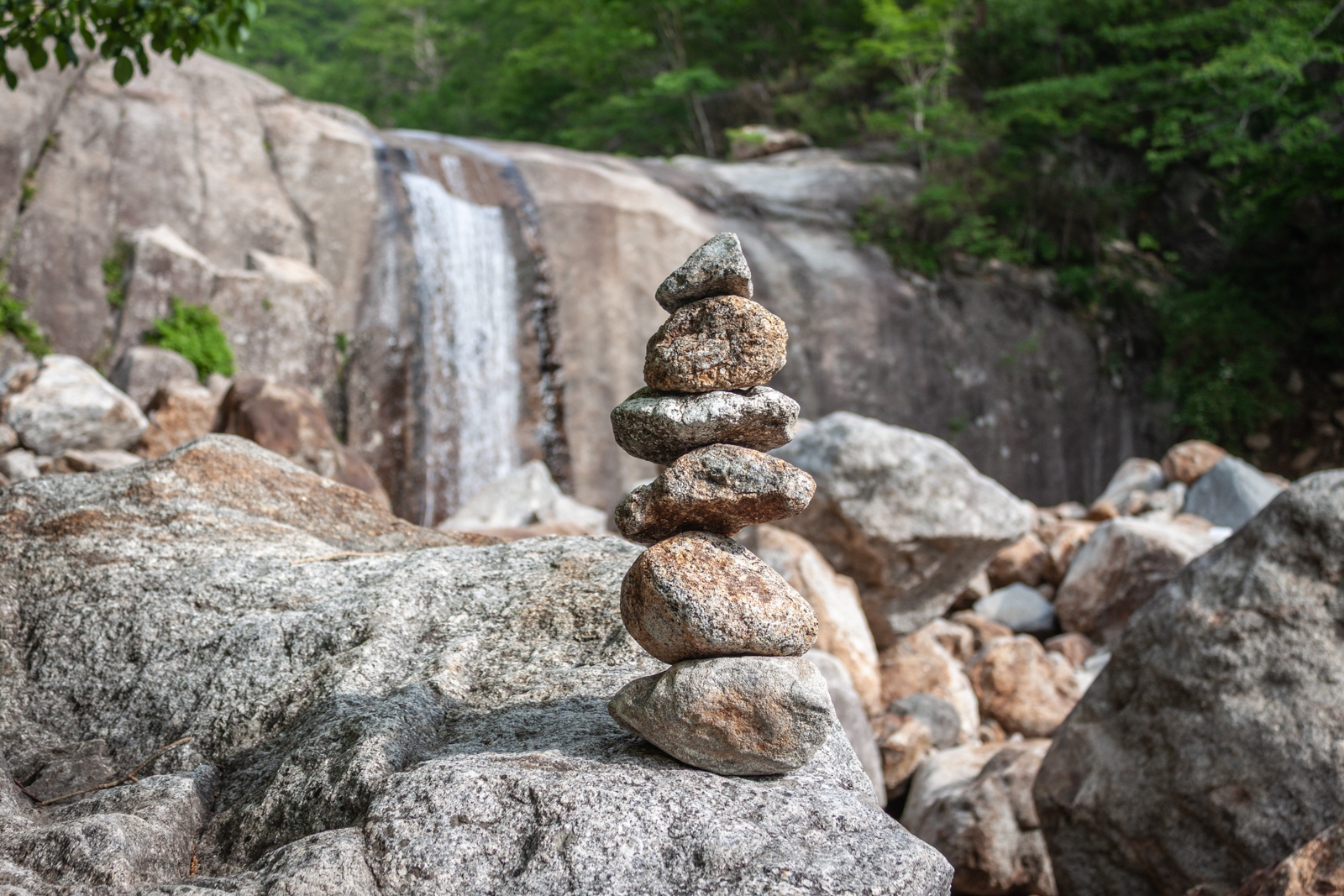 A small, neatly stacked rock cairn on a large boulder in front of a cascading waterfall in a mountainous forest setting.