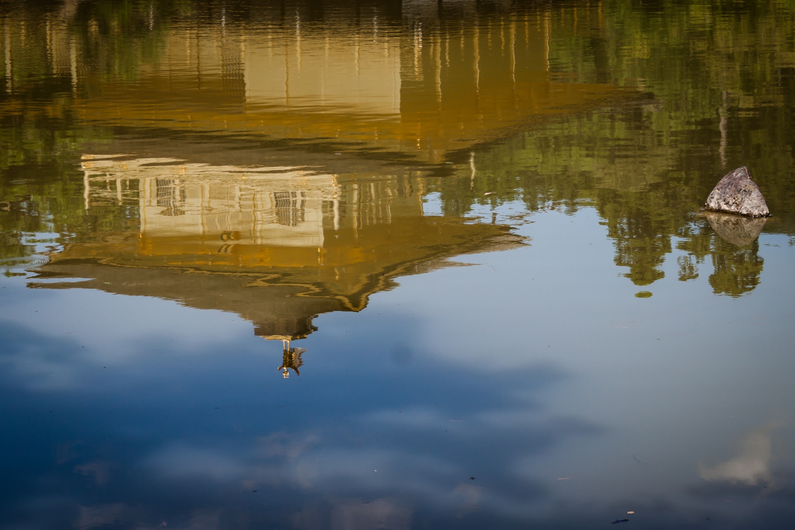 The reflection of a golden pavilion building on the surface of a still pond, with slight ripples distorting the reflection. A single rock juts out from the water, adding to the tranquil and contemplative scene.