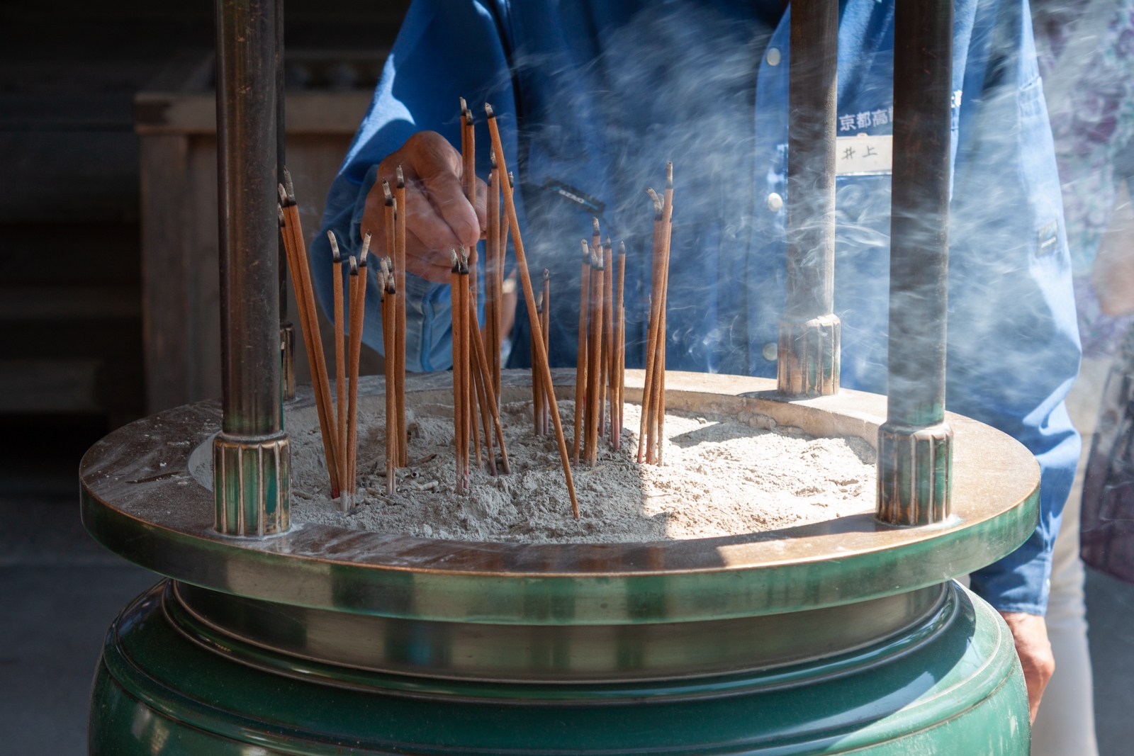 A close-up of an incense burner filled with lit incense sticks. A person’s hand is seen reaching toward the incense, which emits thin streams of smoke, creating a calming atmosphere. The metal burner has a green and gold finish.