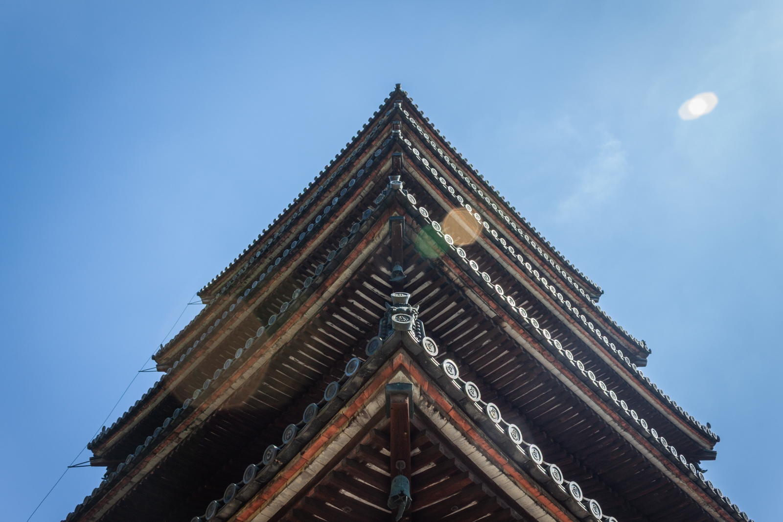 A close-up view of the roof of a Japanese pagoda against a clear blue sky. The intricate design of the roof’s eaves and the decorative tiles create a strong geometric pattern, with light gently illuminating the edges.