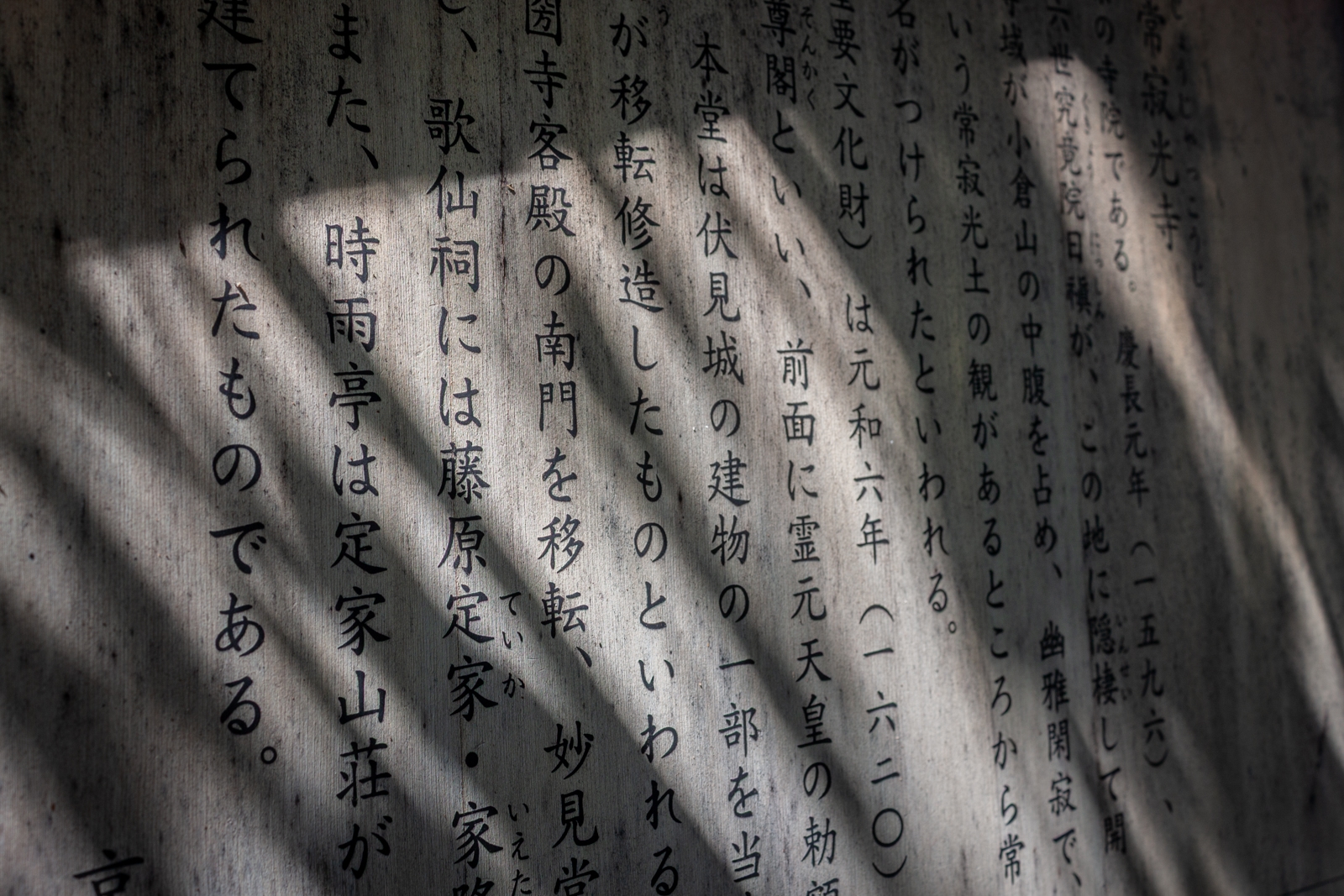 A close-up of Japanese text carved into a stone tablet, with soft shadows of leaves and trees cast upon the surface, giving the image a peaceful and natural atmosphere.