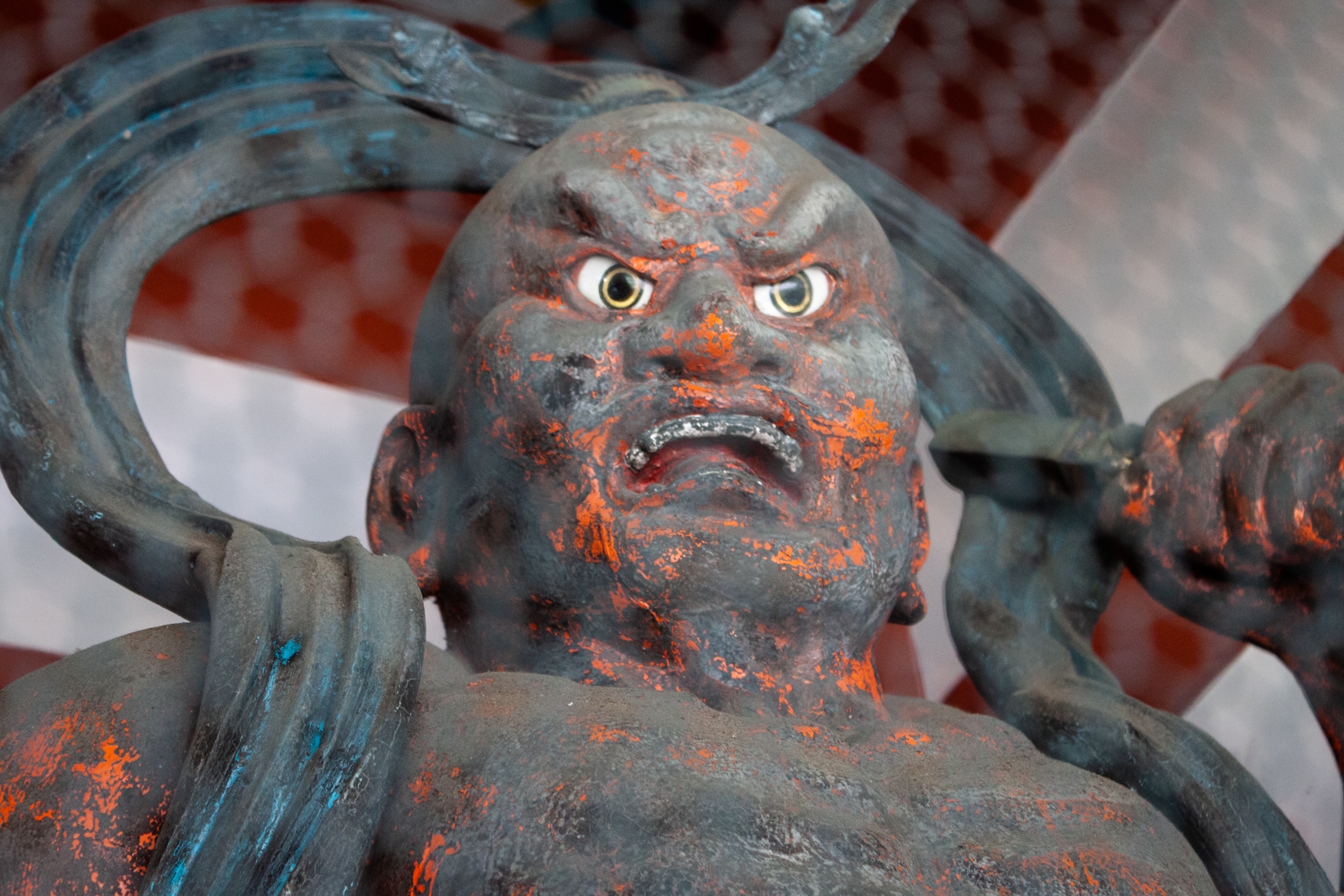 A close-up of a weathered statue of a fierce warrior or guardian figure with an intense expression, gripping a weapon. The statue shows signs of aging with chipped paint and a rough surface, giving it a worn, yet powerful presence.