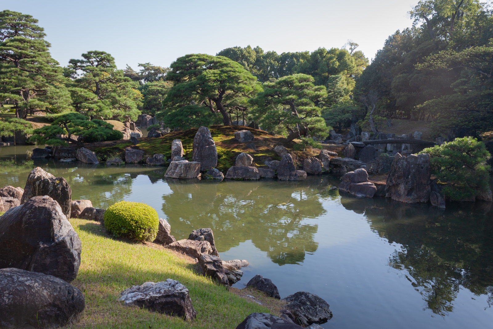 A serene Japanese garden with a large pond surrounded by rocks and lush green trees. Small islands with moss-covered stones and carefully pruned pines reflect in the still water, offering a peaceful and tranquil scene.