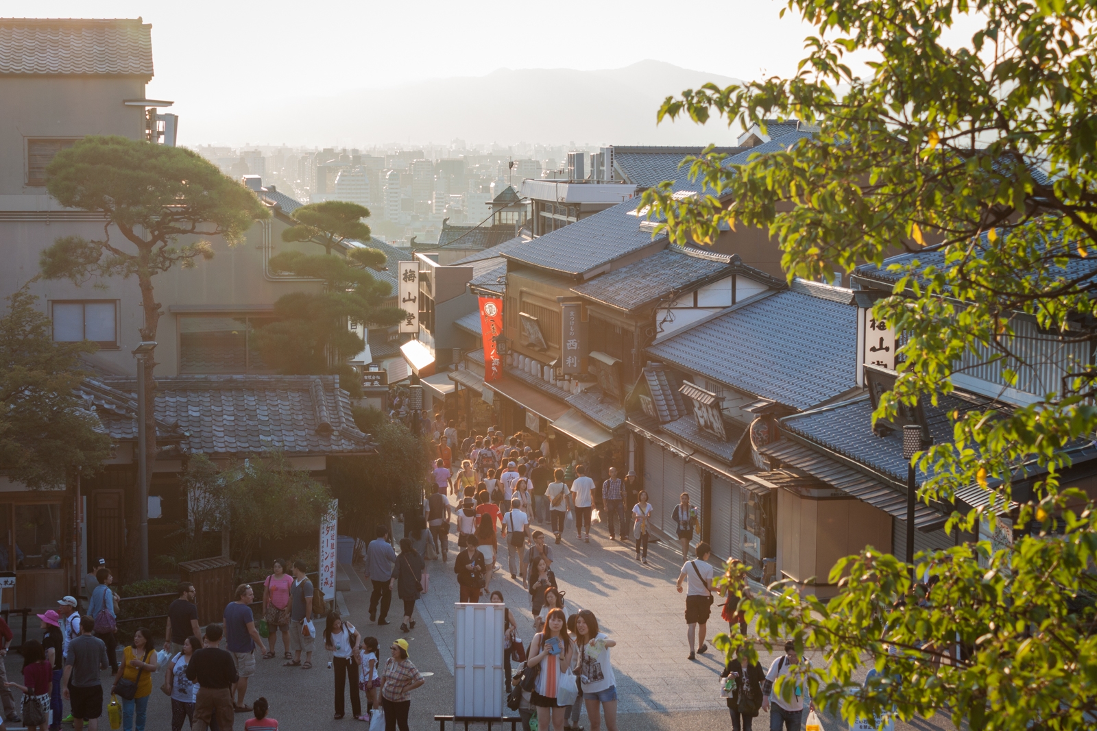 A bustling street filled with people walking between traditional Japanese buildings. The sun sets in the background, casting long shadows and bathing the scene in a warm glow. The street is lined with shops, and trees with green foliage frame the scene.