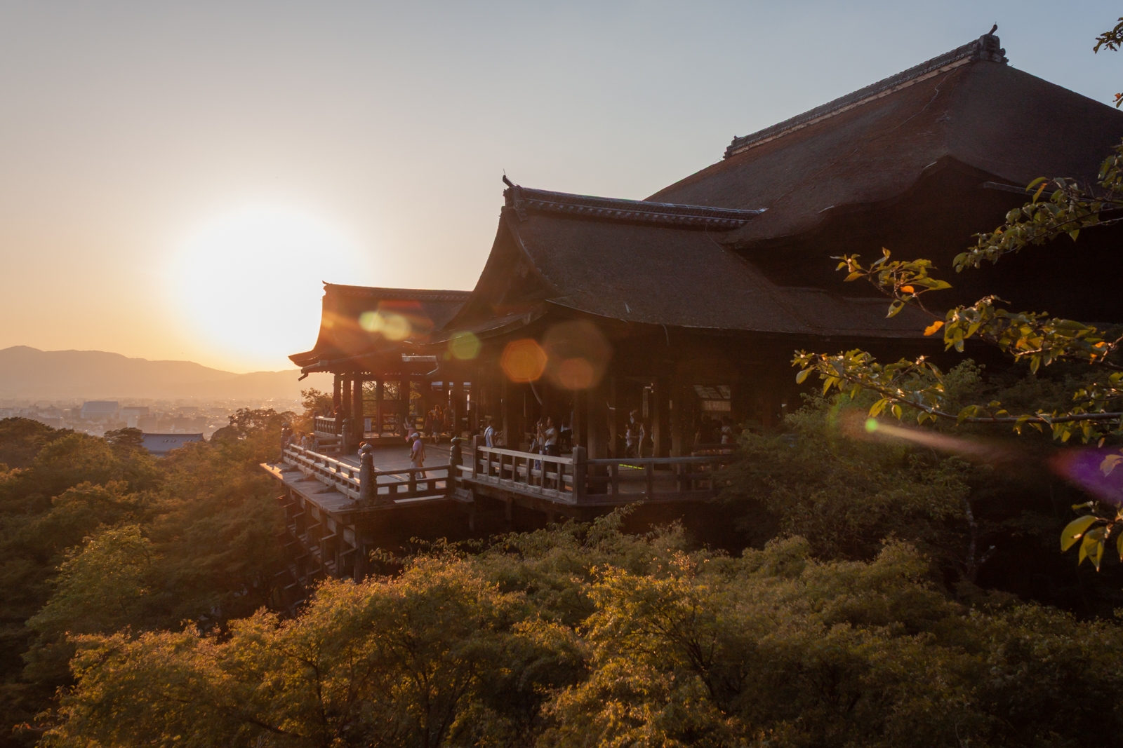 A large wooden temple structure with a balcony overlooking a forested hillside at sunset. The light casts a warm, golden hue across the scene, creating lens flares. People can be seen on the balcony, enjoying the view of the surrounding landscape and distant city.