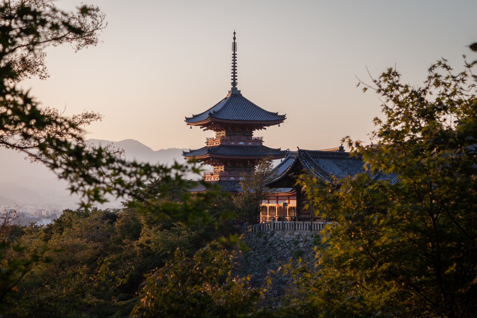 A multi-tiered pagoda rises above a lush forest, with soft light from the setting sun illuminating its intricate roof. In the background, rolling hills and mountains provide a peaceful backdrop, while the pagoda stands in sharp contrast against the natural scenery.