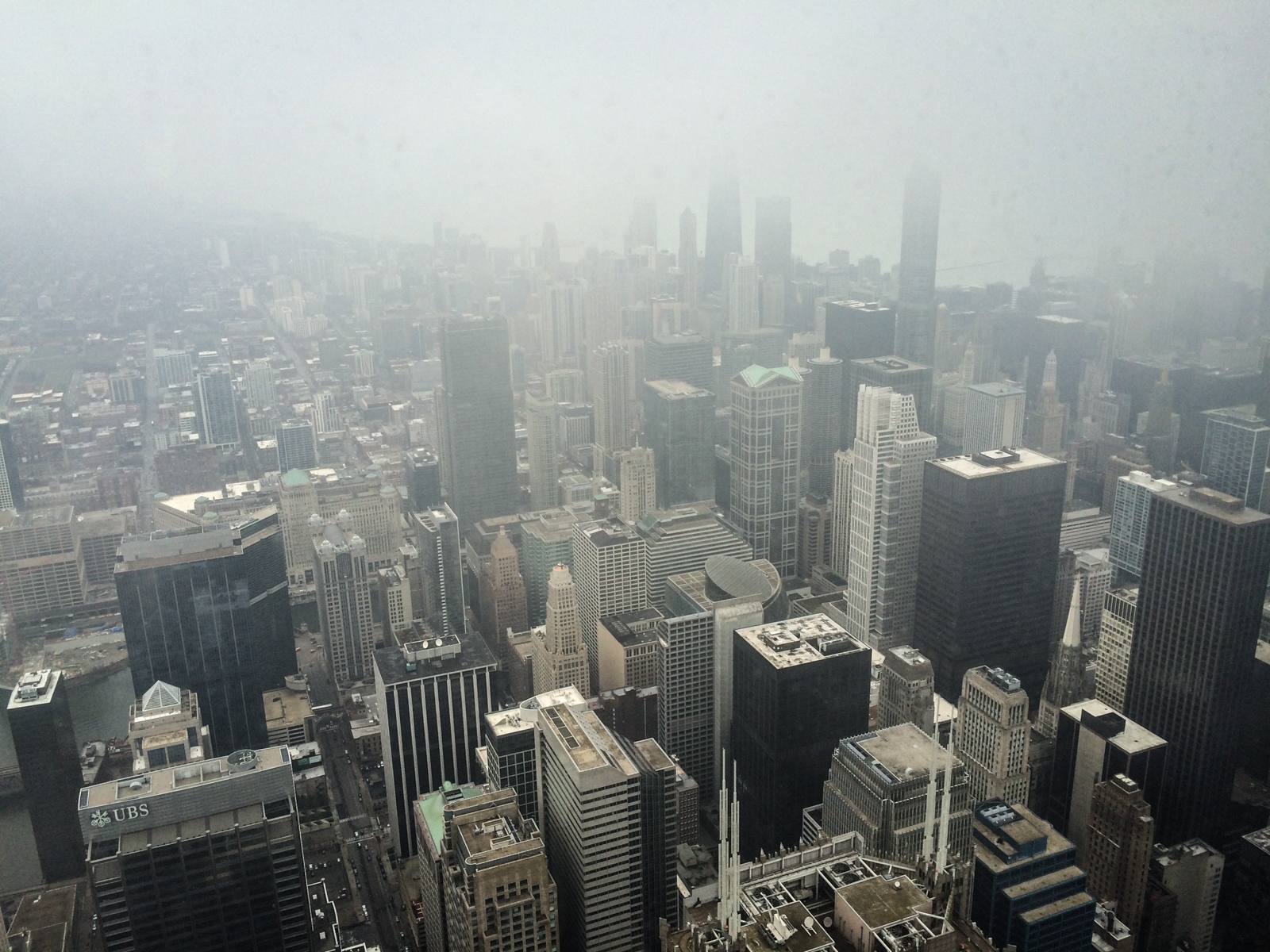 Aerial view of a fog-covered city skyline, with tall skyscrapers stretching out into the distance. The tops of the buildings and parts of the landscape are obscured by thick fog, creating a hazy, atmospheric scene.
