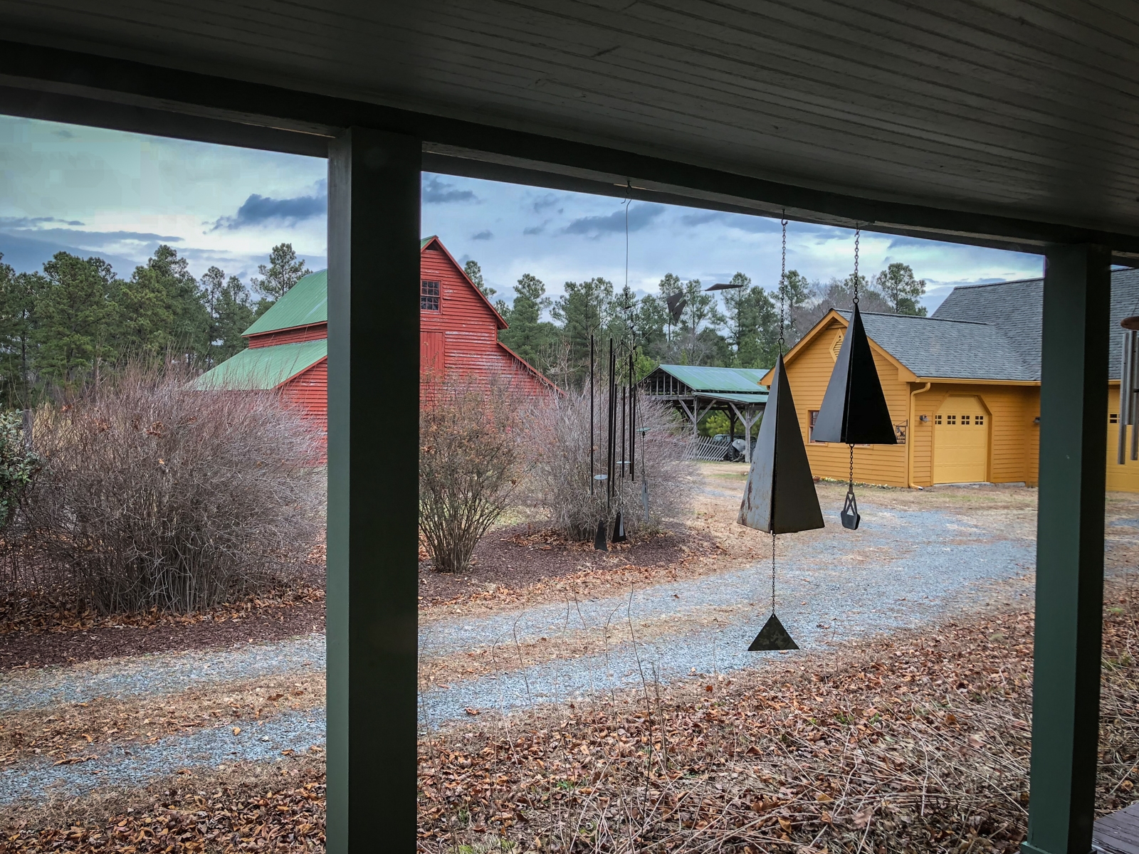 View from a covered porch looking out at a rural property with two brightly colored barns, one red and the other yellow, with green roofs. Wind chimes hang from the porch ceiling, and the surrounding landscape includes gravel paths, bare bushes, and a backdrop of trees beneath a cloudy sky.