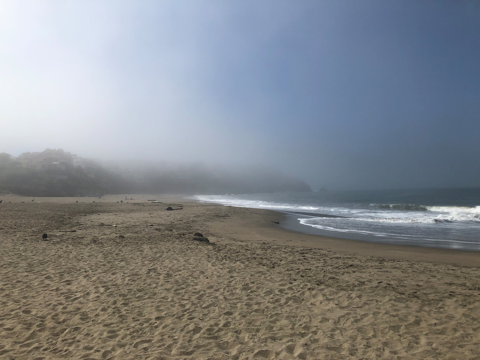 A foggy beach with soft waves rolling onto the sandy shore. The sky and horizon are obscured by thick mist, giving the scene a peaceful, almost dreamlike quality. In the distance, faint outlines of houses on a hill can barely be seen through the fog.