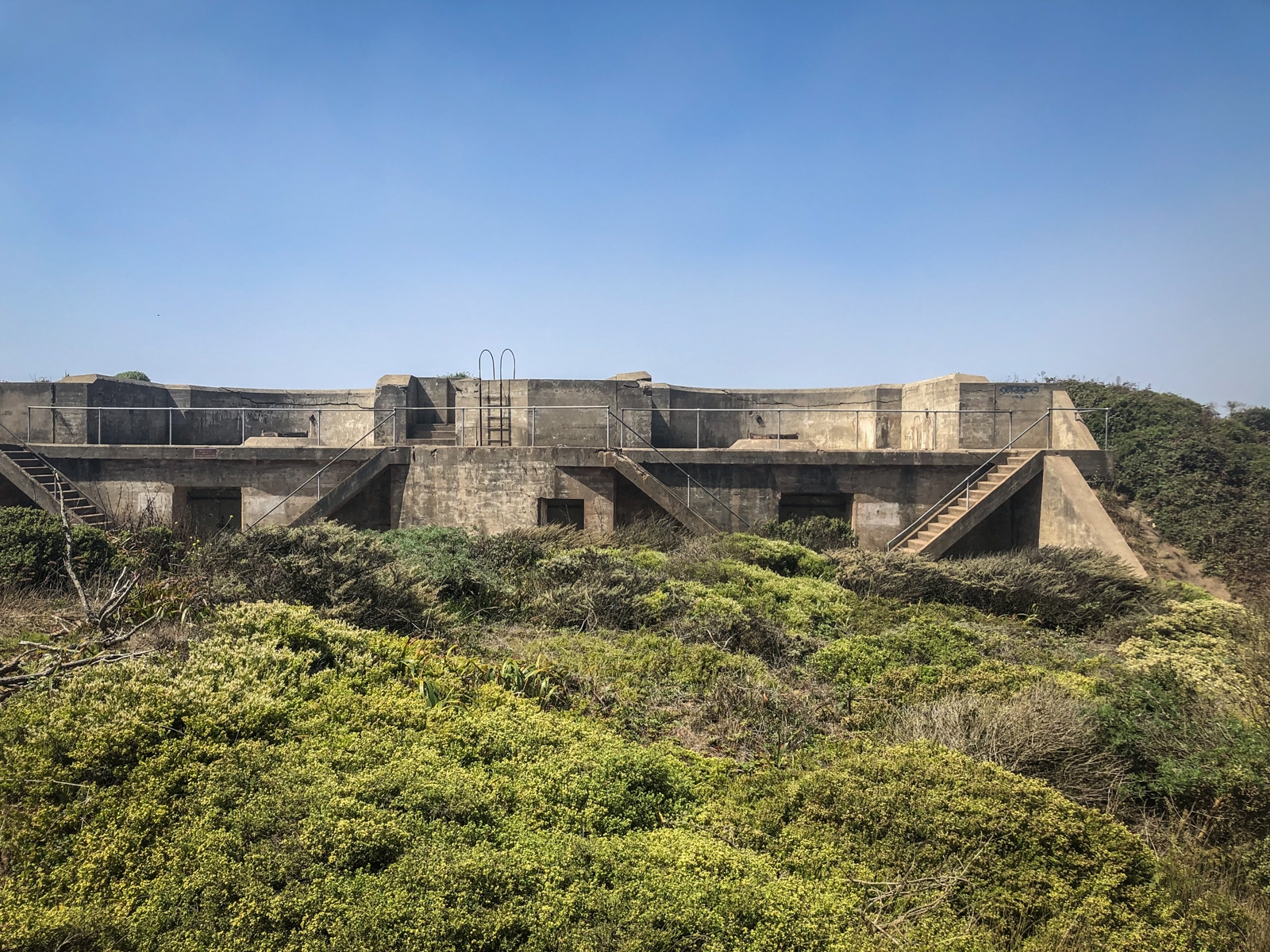 A concrete military bunker structure surrounded by low shrubs and greenery on a sunny day. The structure has stairs leading up to a flat roof with railings and a ladder. The scene evokes a contrast between the harsh, industrial nature of the bunker and the soft, natural landscape around it.