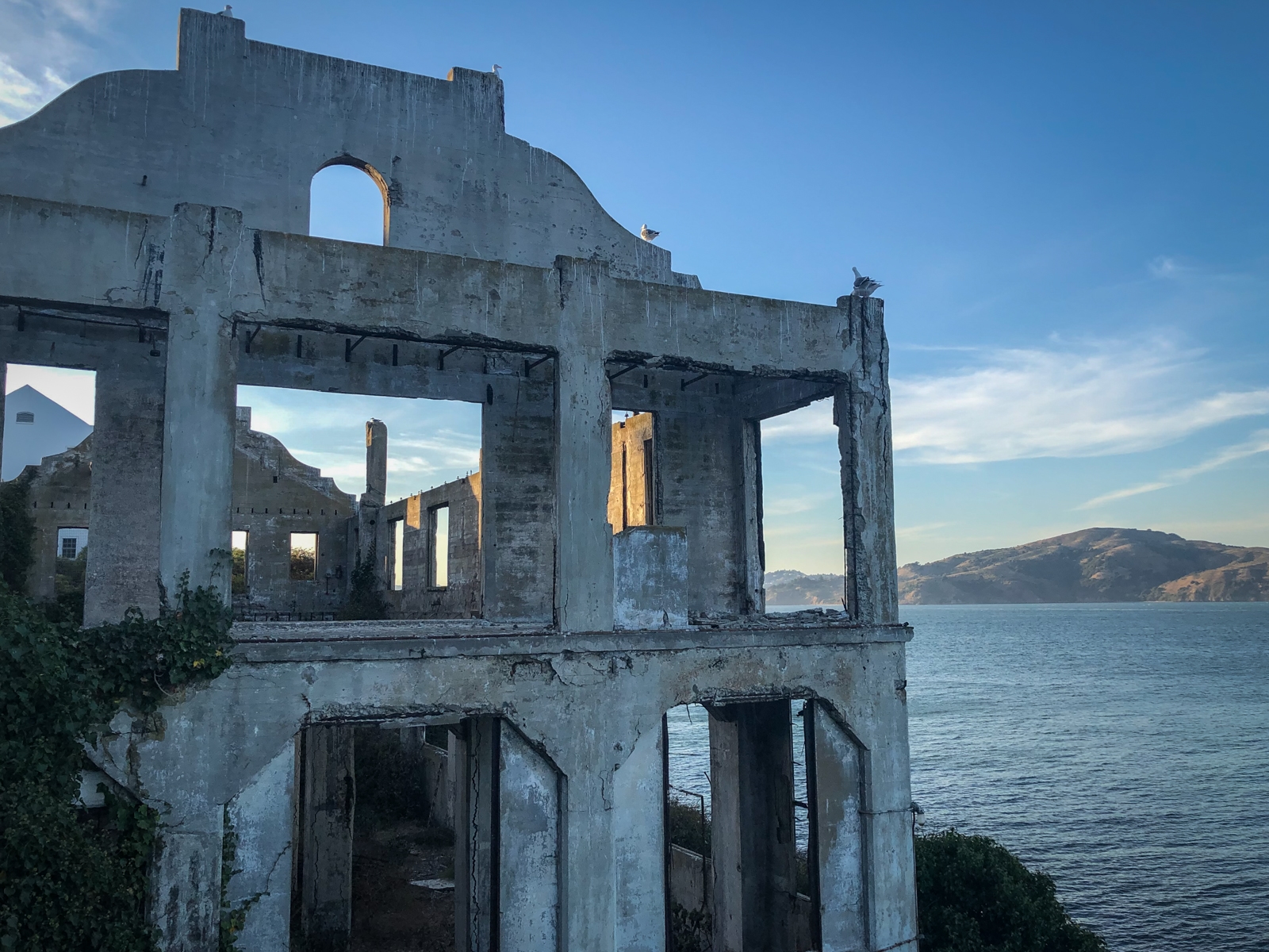 An abandoned, dilapidated concrete structure standing near a calm body of water under a bright, clear sky. The ruins of the building are framed by the rugged hills in the background, creating a contrast between the decaying architecture and the natural beauty surrounding it. Sunlight filters through the empty windows, casting long shadows on the weathered walls.
