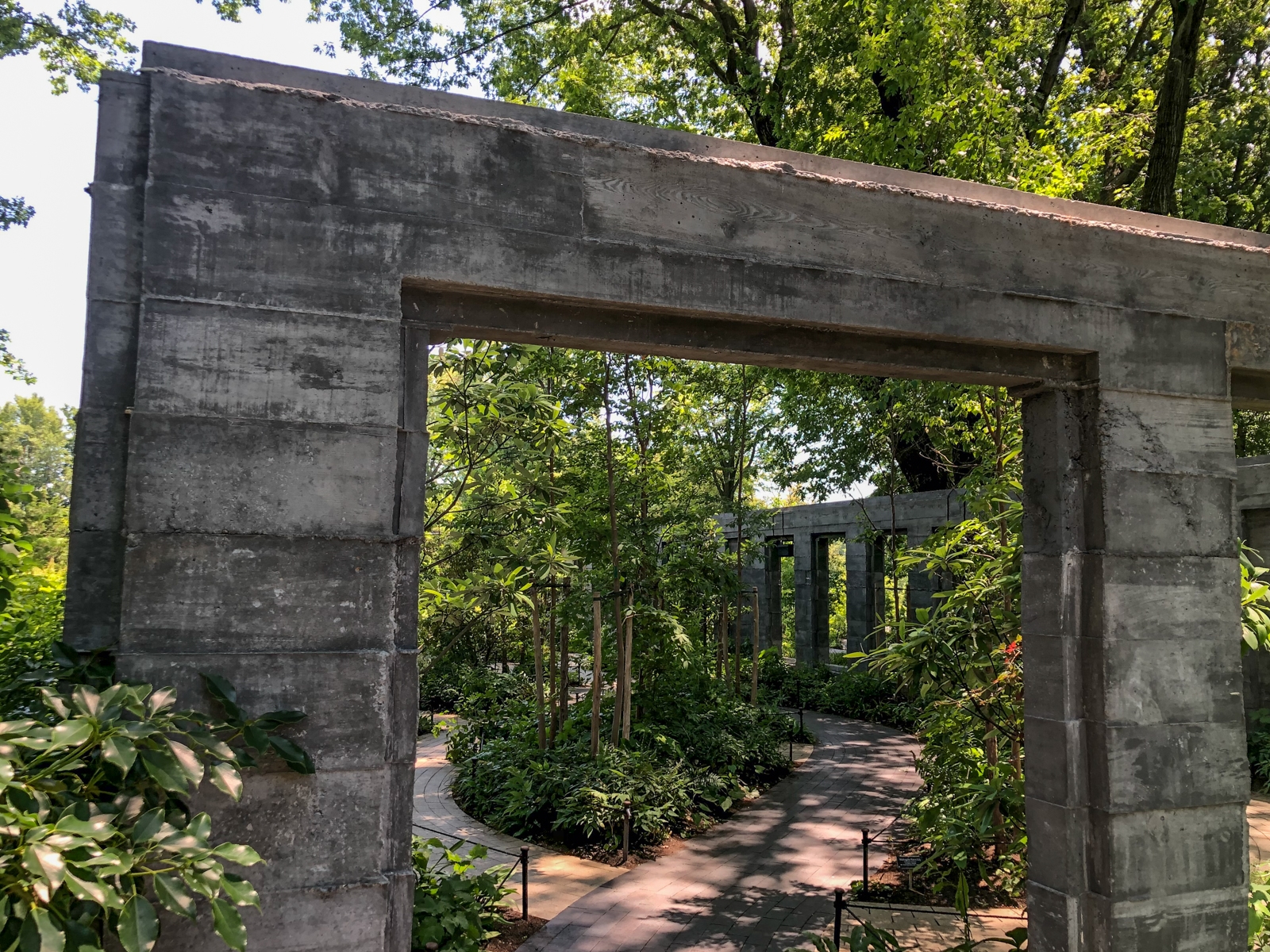 A shaded garden pathway bordered by tall plants and trees, winding through a series of concrete archways. The sunlight filters through the dense foliage, casting soft shadows on the path. The weathered concrete structures add a contrasting modern, architectural element to the natural surroundings.