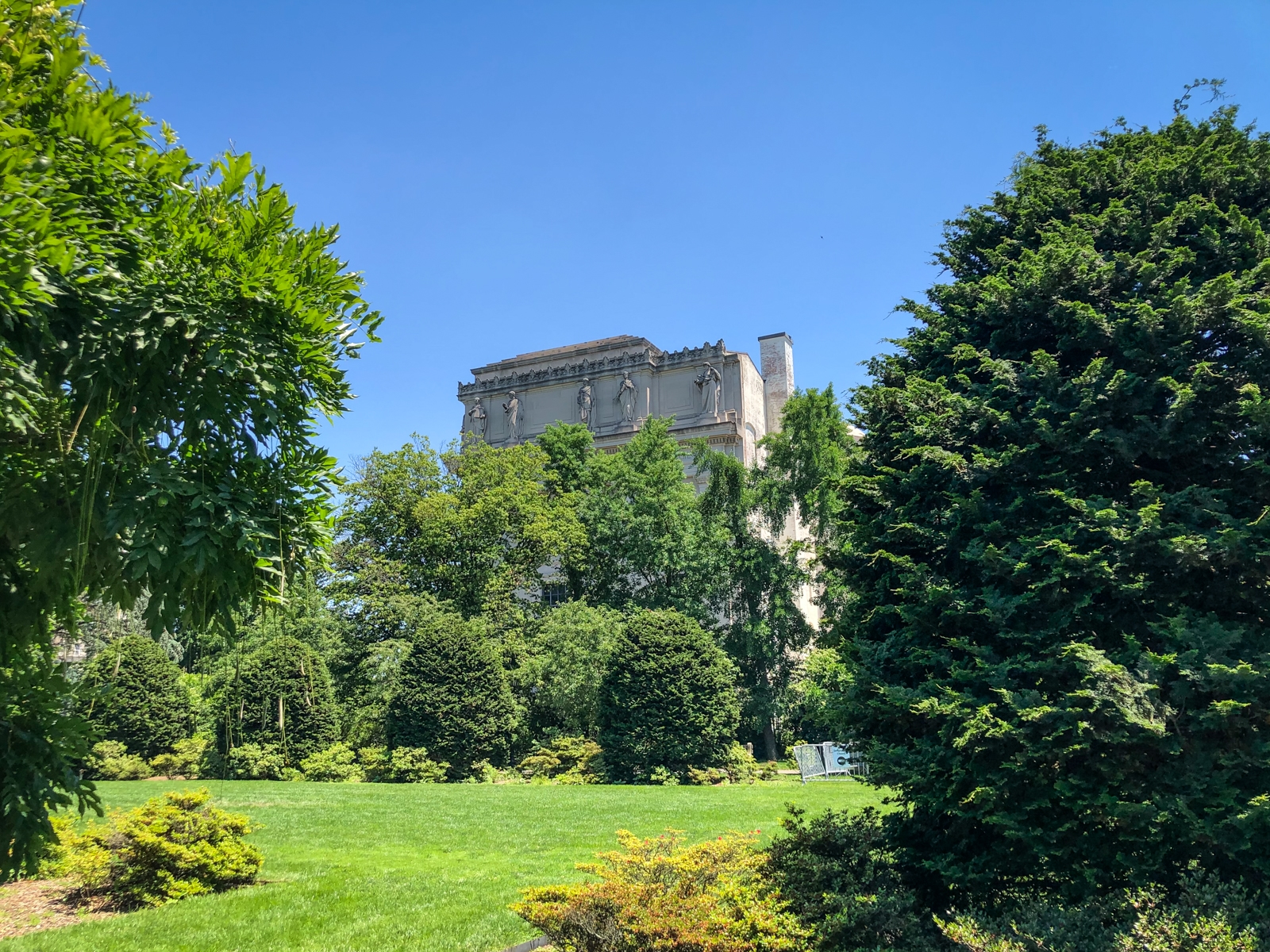 A bright summer day in a garden with manicured green lawns and lush trees. In the background, a classical-style stone building with decorative carvings is partially visible, surrounded by tall greenery. The scene is framed by vibrant trees and shrubs, creating a peaceful, natural setting.