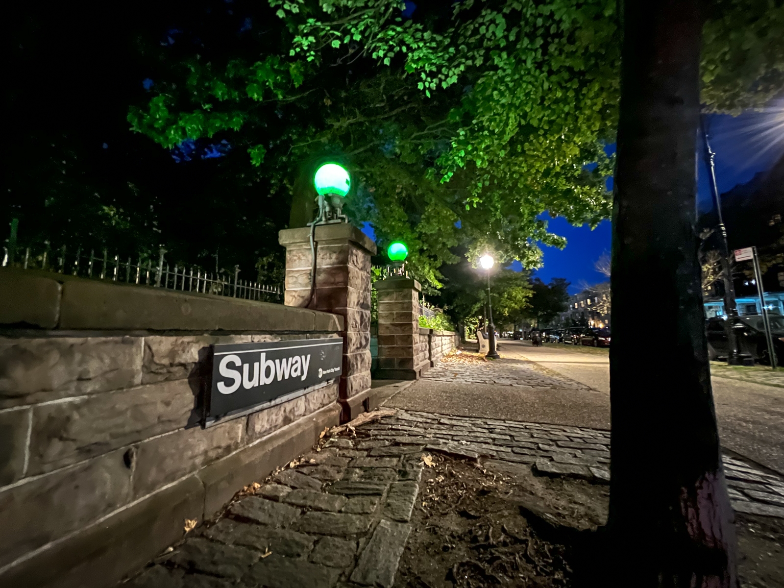 A nighttime street scene with a subway entrance sign set against a low stone wall. Green glowing lamps atop stone pillars and leafy trees line the sidewalk, creating a serene, slightly eerie atmosphere.