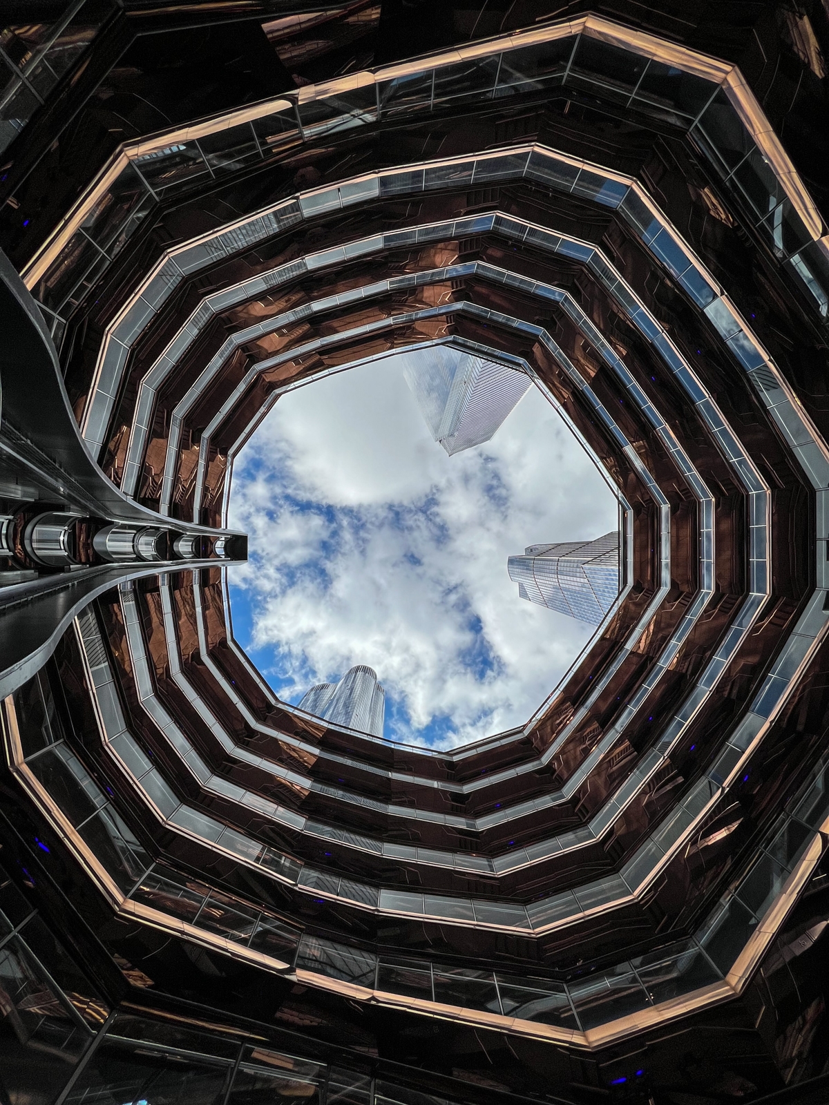 A striking view looking upward through a geometric, multi-level structure with octagonal layers. The open sky above is framed by the copper-colored building, with its reflective surfaces leading the eye toward the blue sky and scattered clouds. The structure creates a sense of depth, with skyscrapers in the distance adding to the urban atmosphere. The contrast between the angular lines of the building and the soft, natural shapes of the clouds makes the scene feel both modern and expansive.