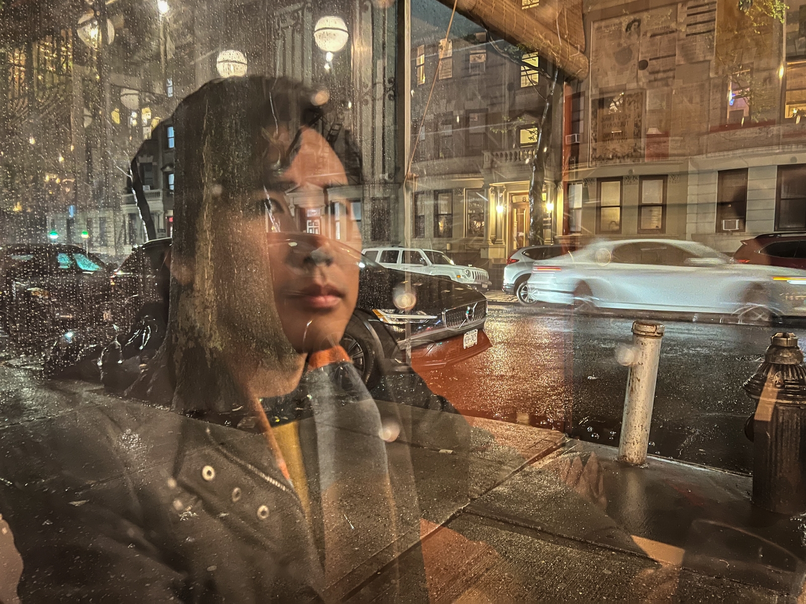 A reflective, moody image of a man seen through a rain-streaked window at night. His face is softly illuminated by the lights outside, while the reflection of city streetlights, cars, and nearby buildings overlay his image. The wet streets and blurred motion of passing cars contribute to the dreamy, layered effect, blending the man's calm expression with the bustling urban scene outside. The image evokes a sense of introspection and solitude amidst the city’s activity.