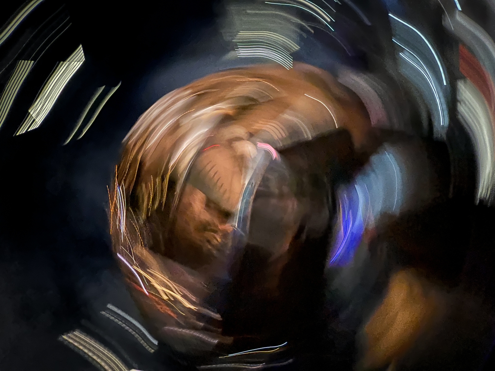 A dynamic, abstract image featuring swirling streaks of light and blurred colors against a dark background. The central focus appears to be a distorted, reflective surface, possibly a metallic object, illuminated by warm and cool tones of light. The circular motion creates a sense of energy and fluidity, giving the impression of rapid movement or a vortex-like effect, while the surrounding lights add a futuristic, dreamlike quality.