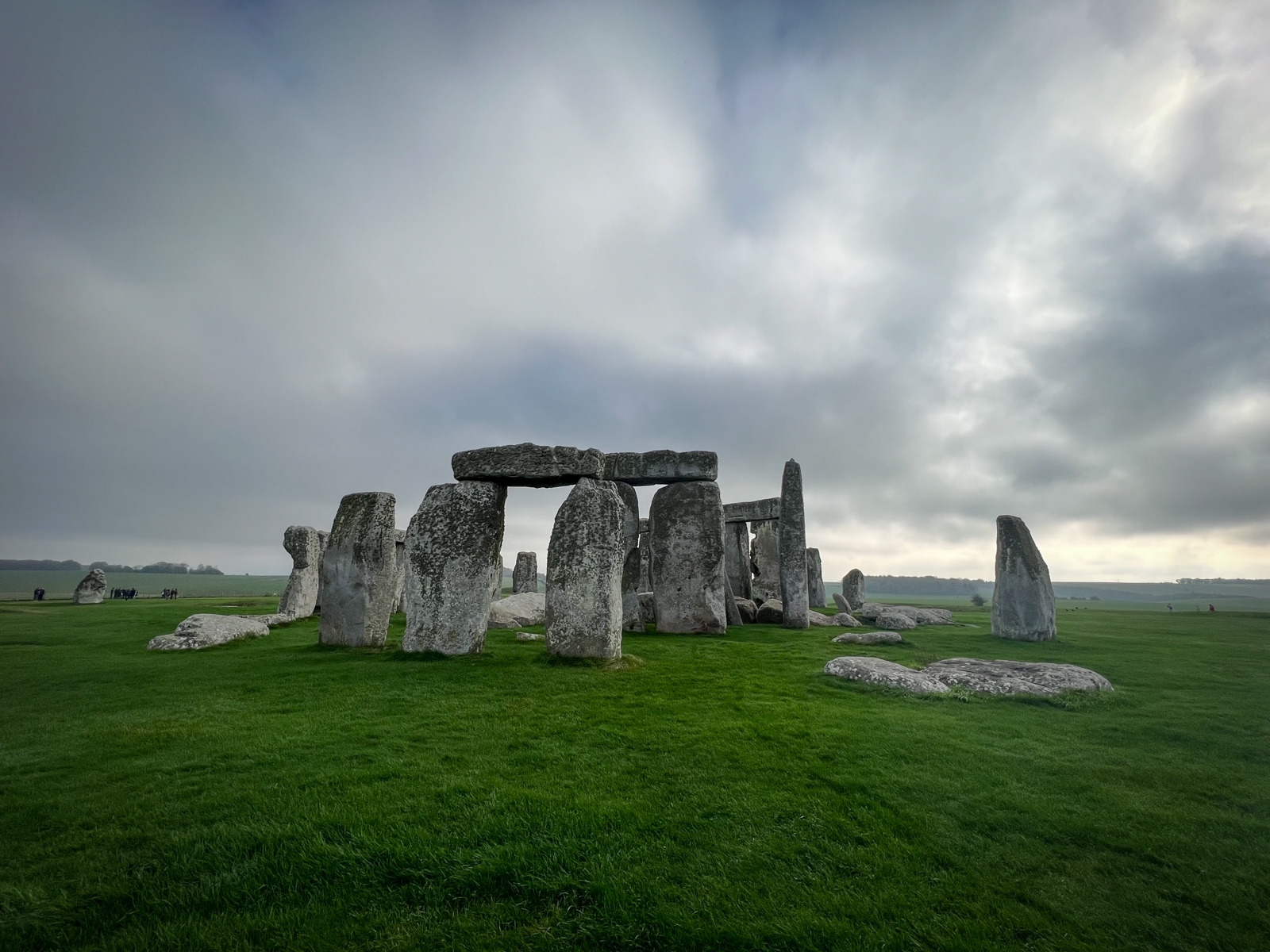 A wide-angle photograph of Stonehenge, the ancient stone monument, under a cloudy, overcast sky. The massive standing stones form a partial circle, with large horizontal lintels balanced on top of vertical stones. The surrounding landscape is a vast, green field, with a few distant figures scattered around, providing a sense of scale. The moody sky adds a dramatic atmosphere, evoking the mystery and timelessness of the site.