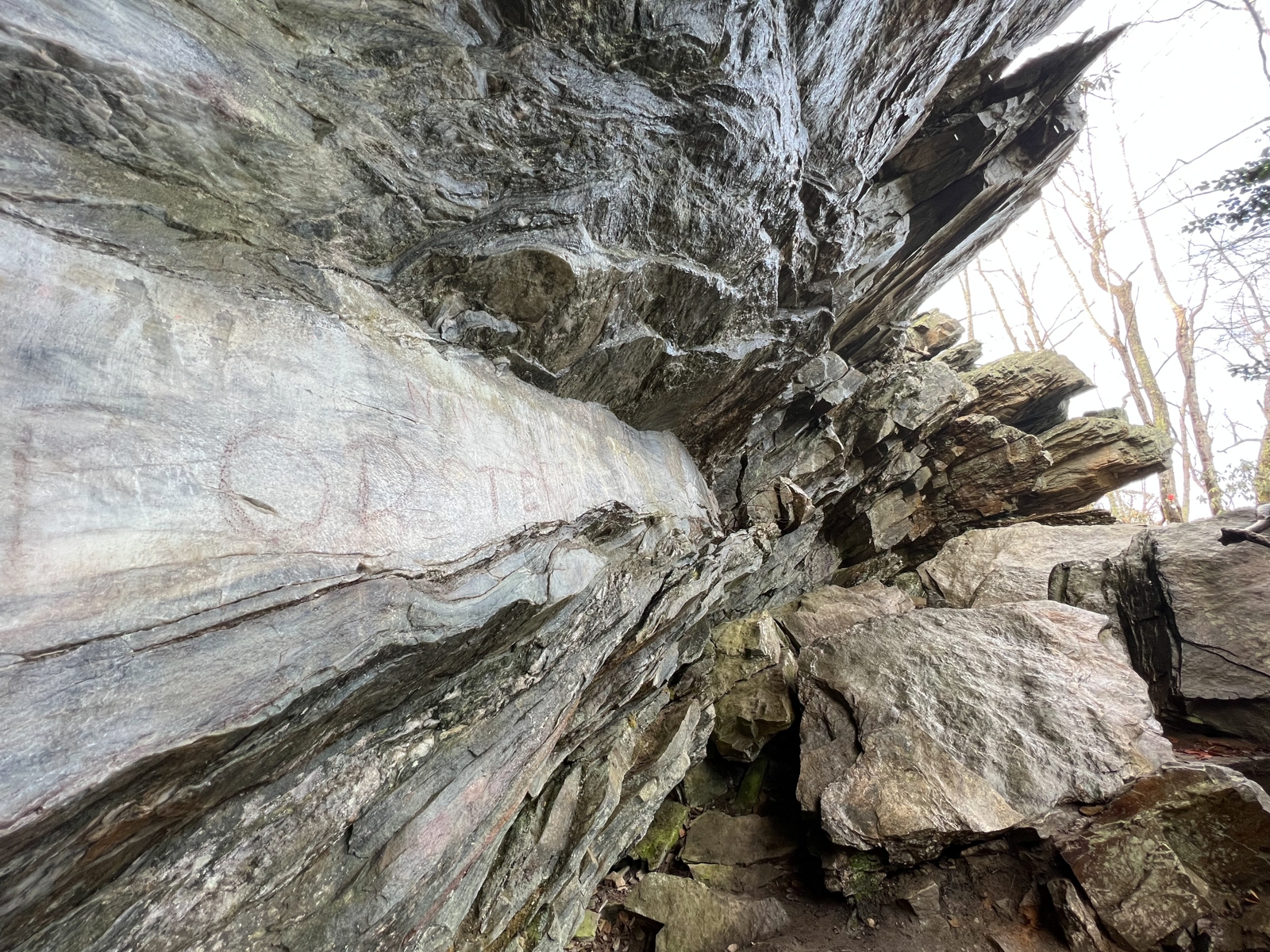 A rugged rock face with layers of stone slanting sharply, leading into a tight crevice. The rock surface is streaked with various earthy tones, and the texture appears weathered and jagged. Bare trees are visible in the background against a pale sky.