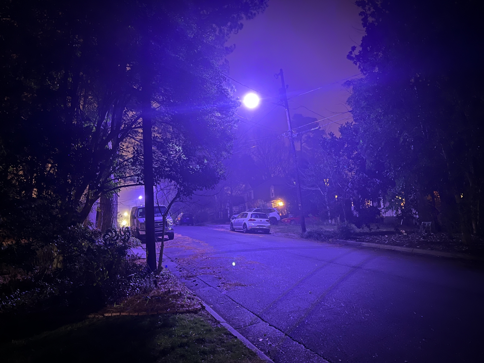A quiet suburban street at night, illuminated by a bright streetlamp casting a purple-hued glow over the scene. The soft mist in the air amplifies the color, creating an otherworldly atmosphere. Cars are parked along the street, and trees with dark silhouettes frame the view. Faint warm lights from houses peek through the branches in the background, contrasting with the eerie blue-purple light.