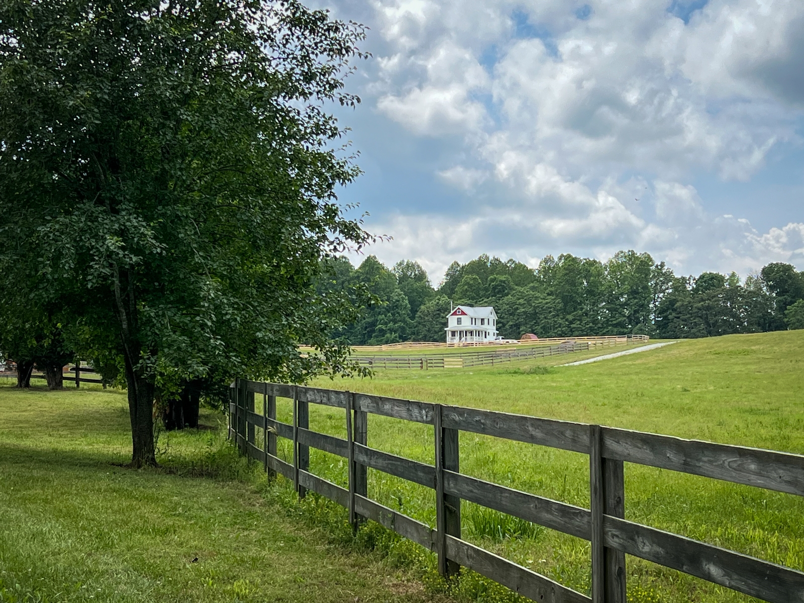 This scenic image features a pastoral landscape with a wooden fence leading the viewer’s eye to a white farmhouse in the distance. The field is lush and green, and the house is surrounded by trees under a partly cloudy sky, evoking a sense of quiet rural life.