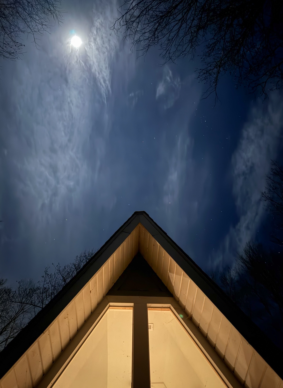 The peaked roof of a house lit warmly from below contrasts with a dark blue night sky, where thin clouds drift past a bright moon and scattered stars.