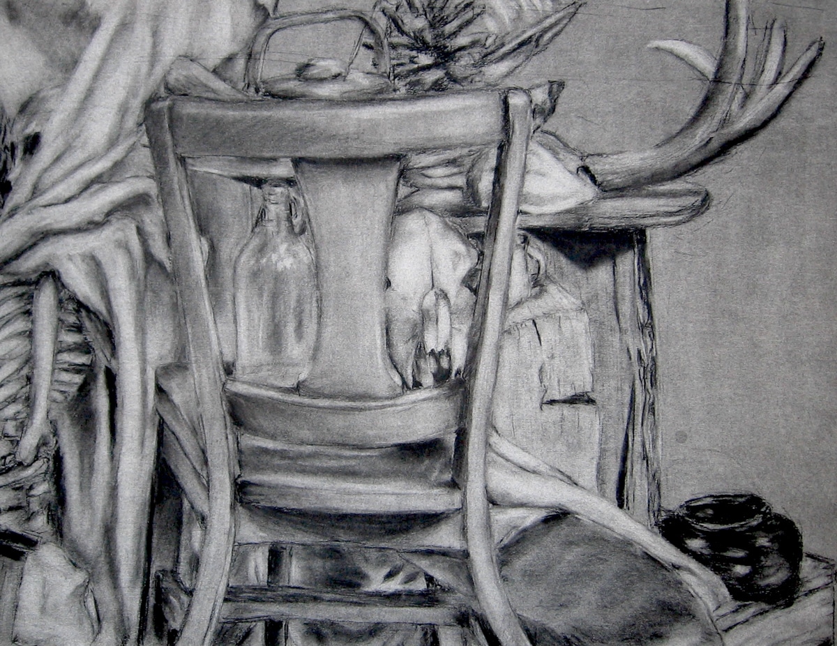 A charcoal still-life drawing featuring a wooden chair in the foreground. Behind the chair, a bottle and an animal skull are visible, creating a rustic and mysterious atmosphere. Antlers extend from the upper right side, adding to the natural and slightly eerie setting. The composition includes draped fabric and other objects, blending textures and forms in a detailed, layered arrangement.